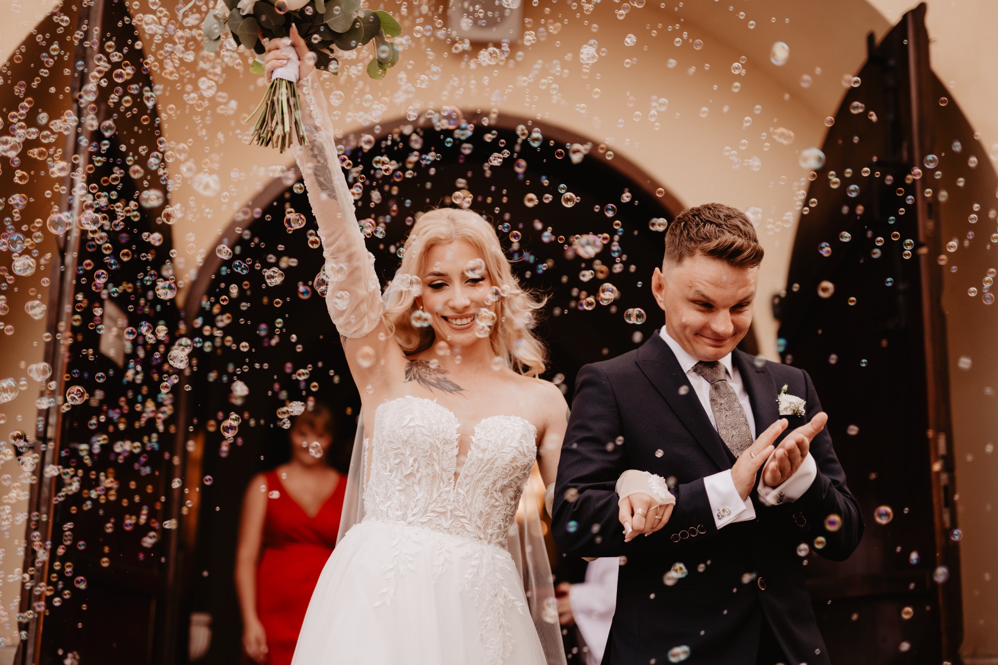 Uśmiechnięta para młoda wychodzi z kościoła, otoczona bańkami mydlanymi. Panna młoda w sukni z koronką, pan młody w ciemnym garniturze. W tle widoczna kobieta w czerwonej sukience.