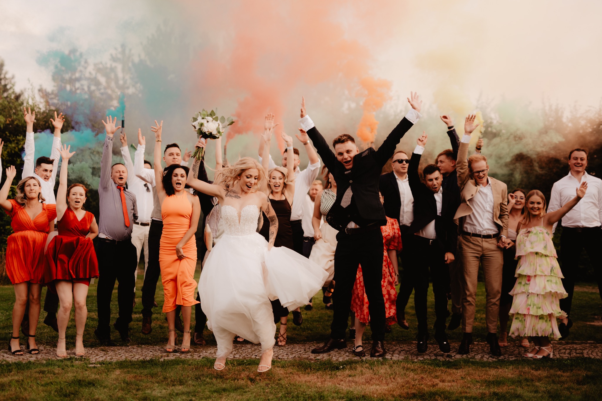 Radosny skok młodej pary i gości weselnych z kolorowymi dymami w tle. Panna młoda w zwiewnej sukni, pan młody w garniturze, wszyscy z podniesionymi rękami świętują.