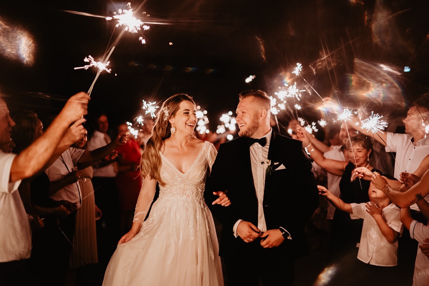 Uśmiechnięta para młoda w otoczeniu gości weselnych z zimnymi ogniami, nocne ujęcie. Panna młoda w sukni z długim rękawem, pan młody w czarnym smokingu.