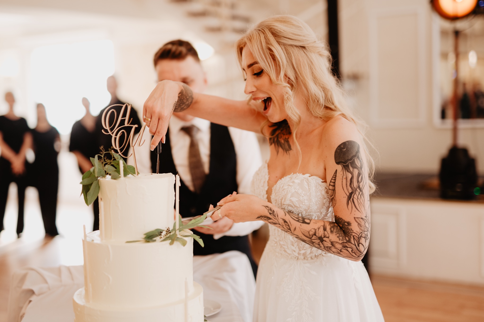 Panna młoda z tatuażami kroi tort weselny, obok pan młody. W tle goście i jasne wnętrze sali. Dekoracja tortu z inicjałami. Uśmiech i radość.