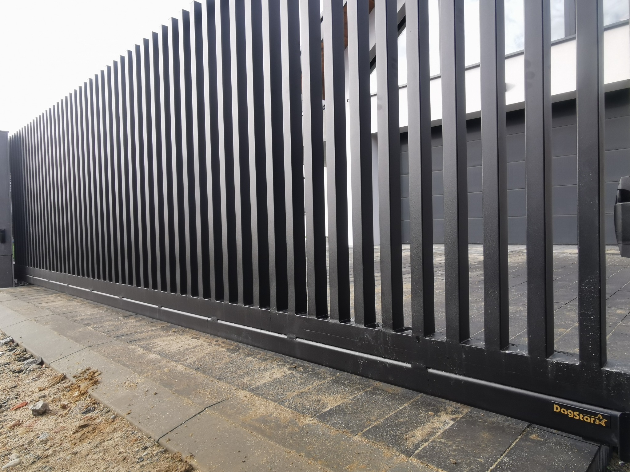 Nowoczesna, przesuwna brama metalowa z pionowymi szczeblami w kolorze grafitowym. Widoczne detale konstrukcyjne i fragment posesji w tle.