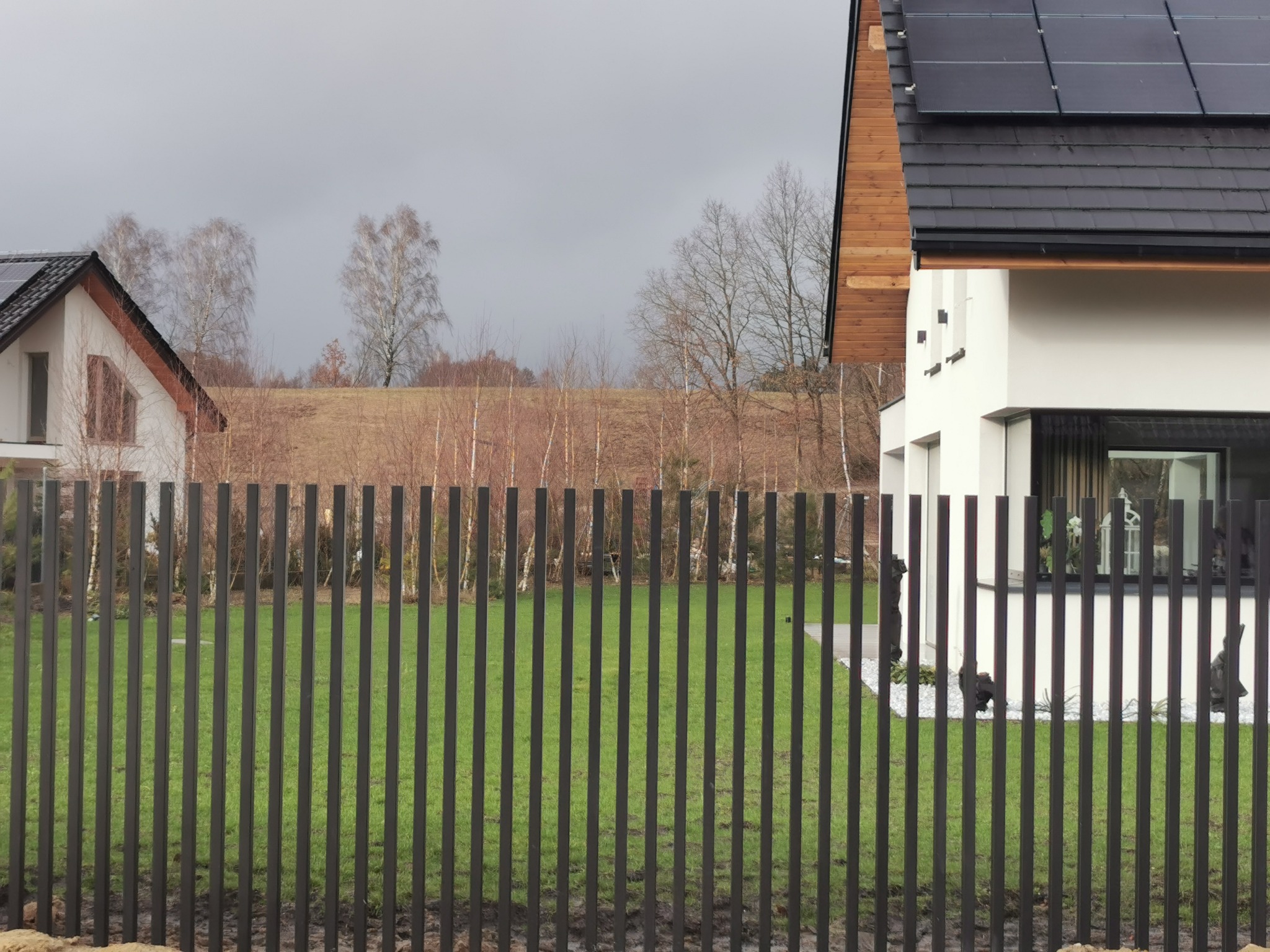 Nowoczesne ogrodzenie palisadowe w kolorze antracytu, na tle domu z panelami słonecznymi i trawnika. Minimalistyczny design i stonowana kolorystyka.