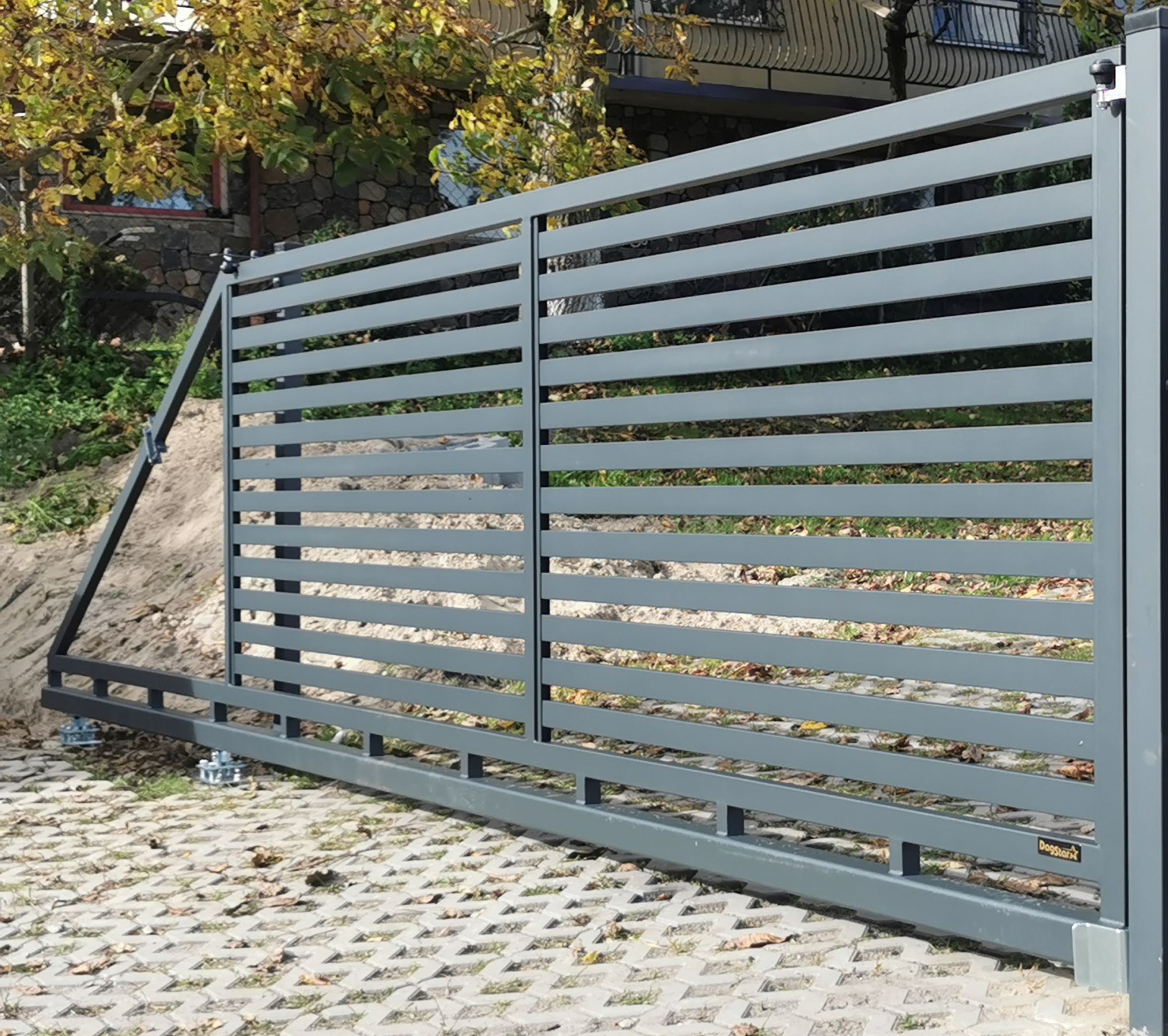 Nowoczesna, przesuwna brama w kolorze grafitowym z poziomymi panelami. Widoczny mechanizm jezdny i fragment posesji z kostką brukową oraz roślinnością.