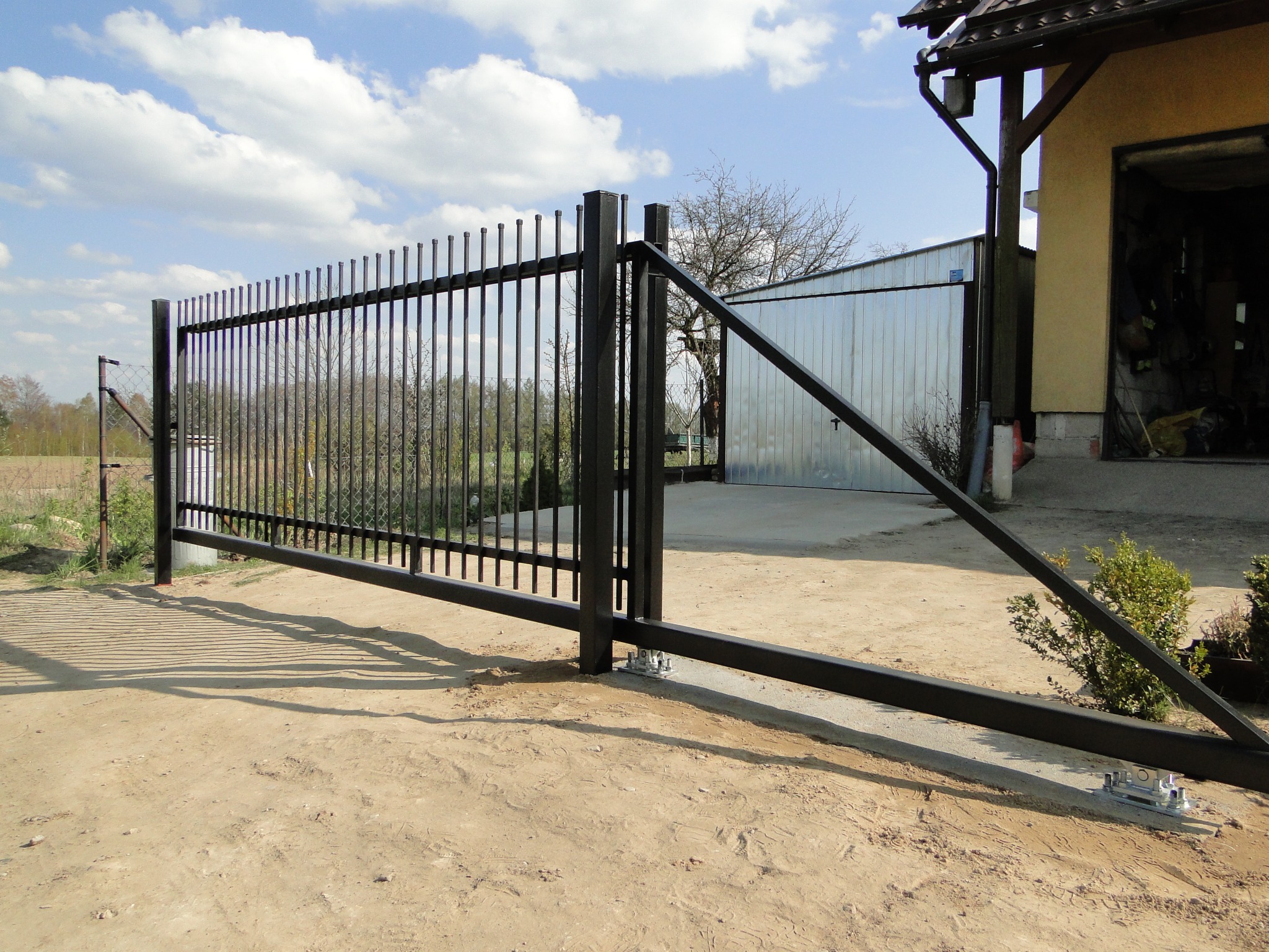 Nowoczesna, przesuwna brama metalowa z pionowymi prętami, zamontowana na betonowej podstawie. W tle budynek gospodarczy i błękitne niebo z chmurami.