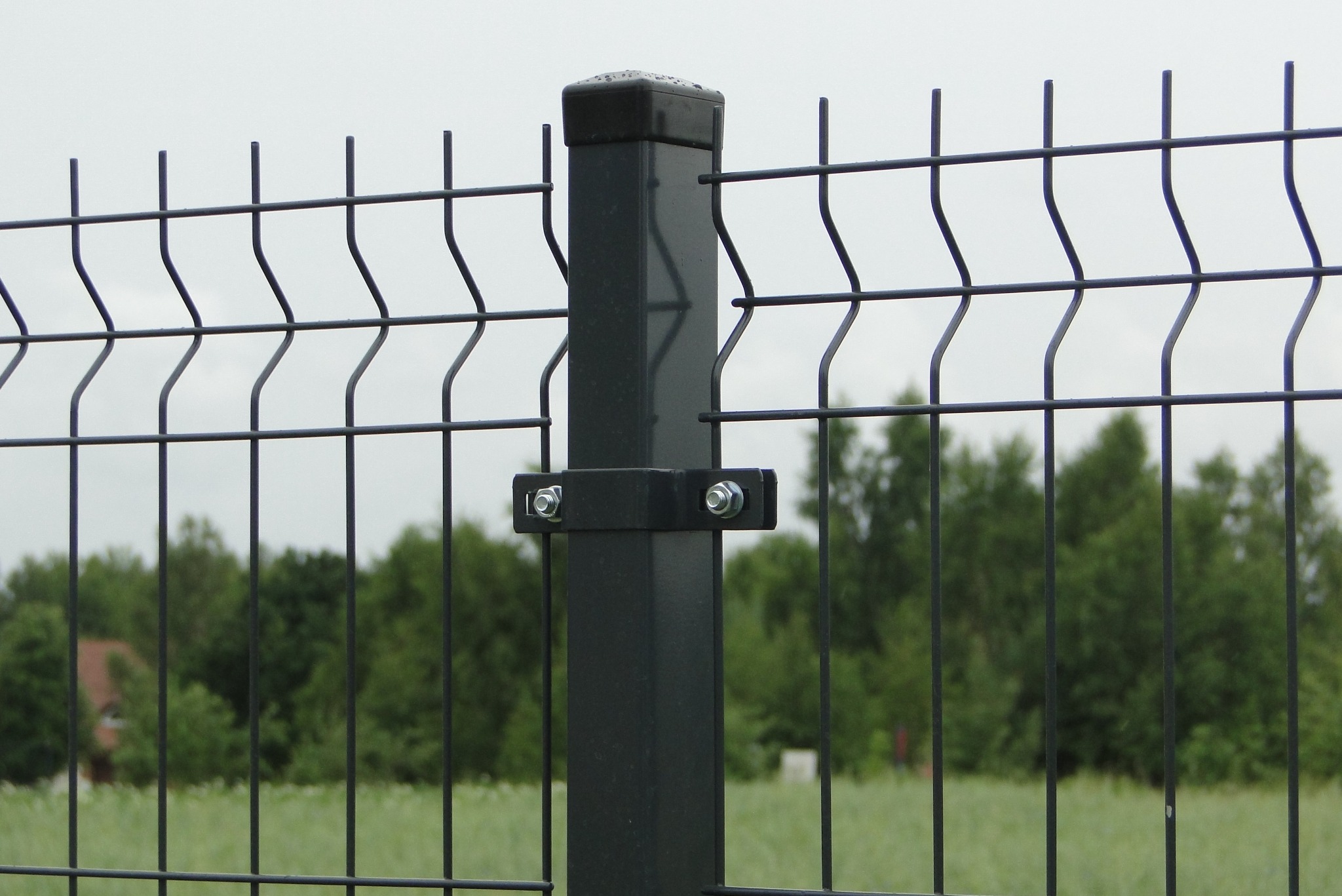 Fragment grafitowego ogrodzenia panelowego z profilem mocującym, na tle zieleni i szarego nieba. Widoczne detale łączeń i mocowań.