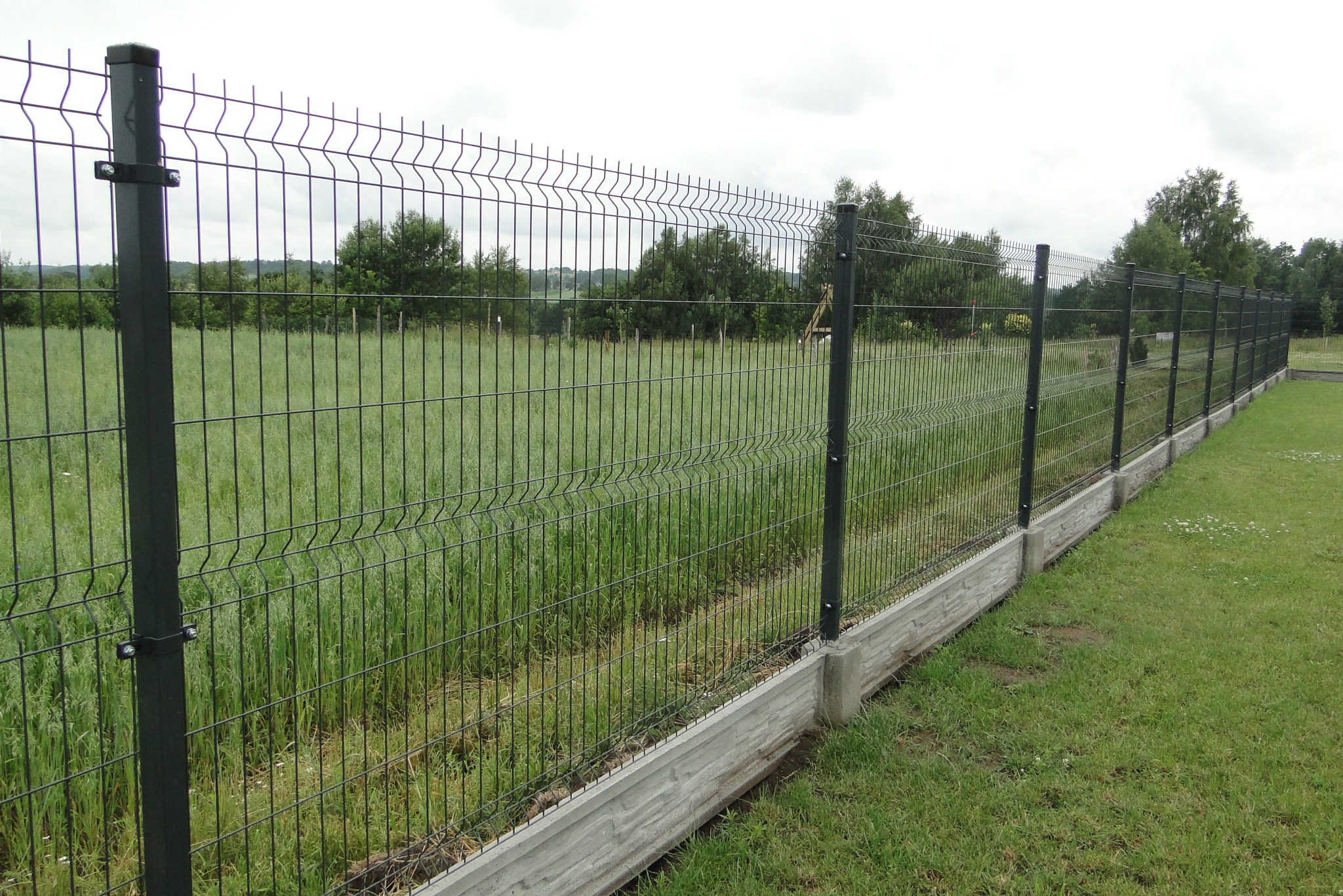Długie, panelowe ogrodzenie w kolorze grafitowym z betonową podmurówką, oddzielające trawnik od łąki z drzewami w tle, w pochmurny dzień.