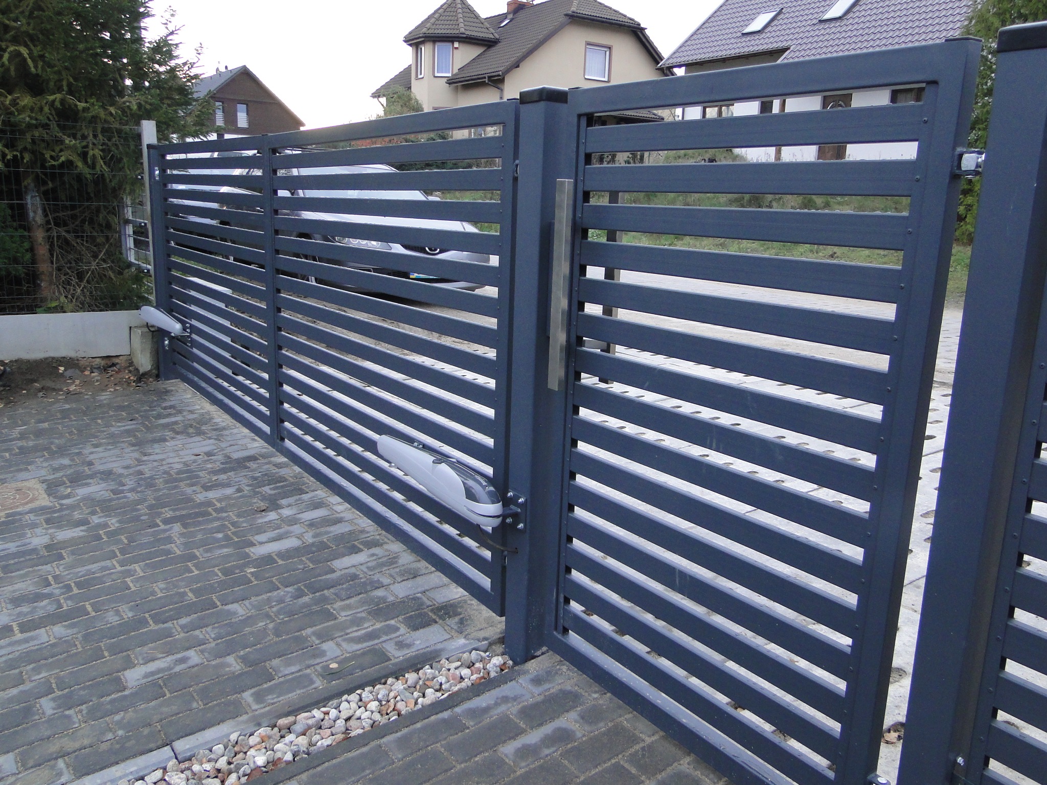 Nowoczesna brama metalowa z poziomymi panelami i automatycznym napędem, na tle brukowanej posesji z widocznym domem w tle.