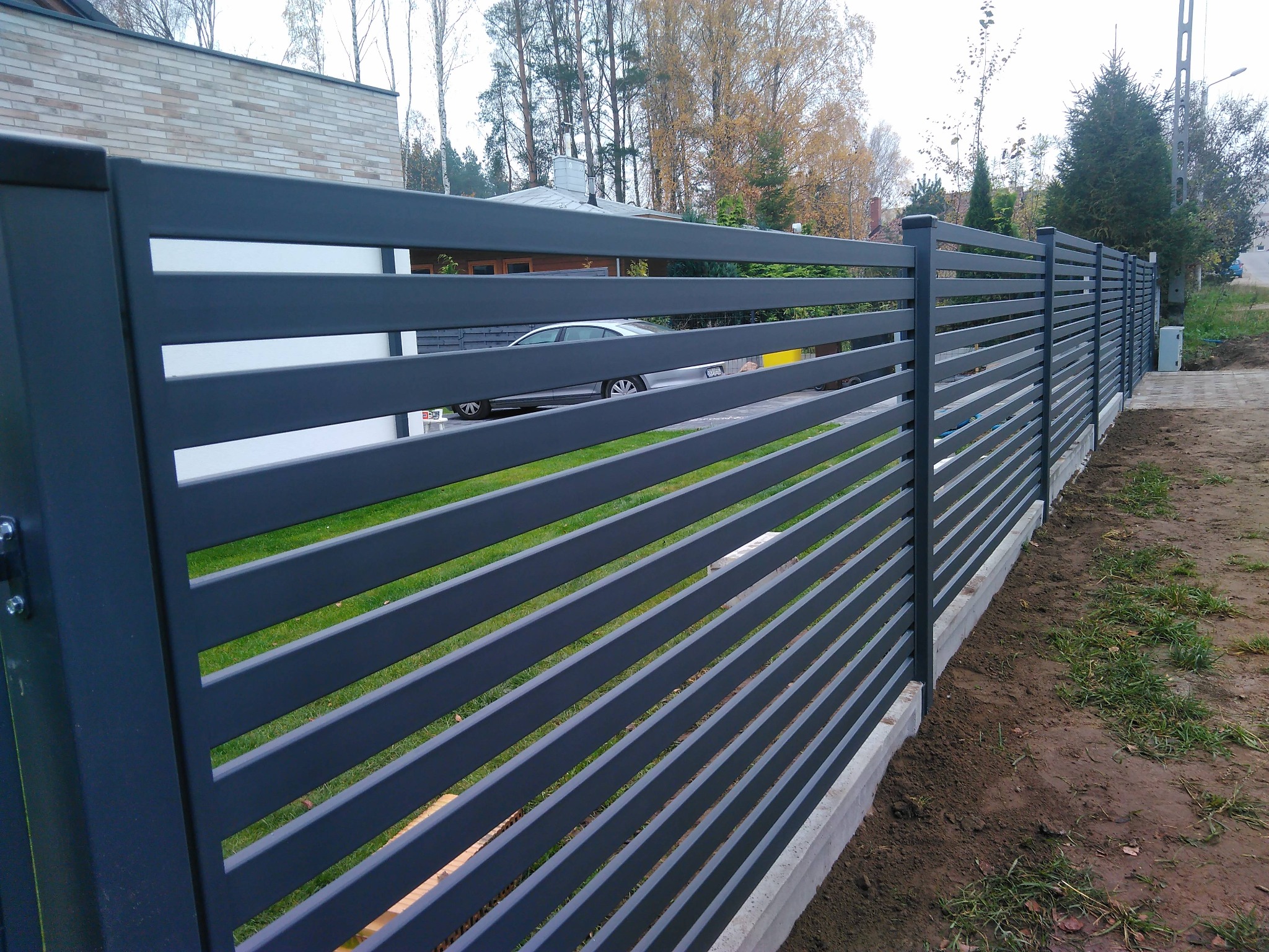 Nowoczesne ogrodzenie metalowe z poziomymi przęsłami w kolorze grafitowym. Widoczne słupki i podmurówka, w tle dom i zieleń. Ujęcie z boku.