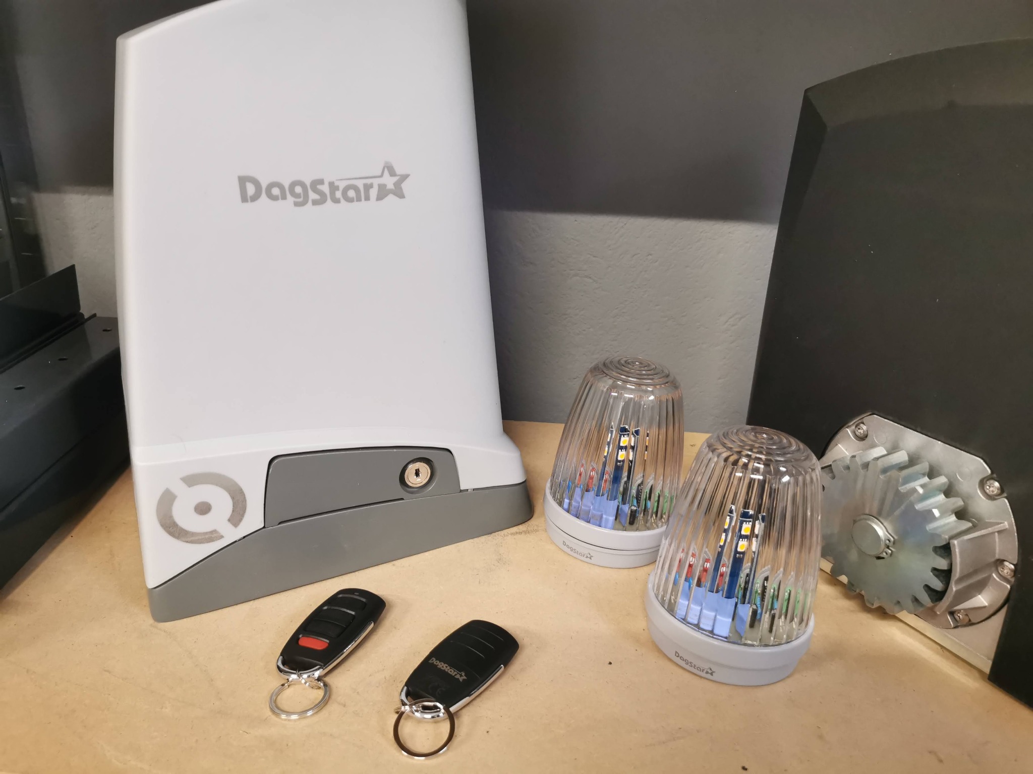 Zestaw do bramy: napęd z logo Dagstar, dwa piloty, lampy ostrzegawcze i przekładnia zębata. Elementy na blacie, gotowe do montażu.