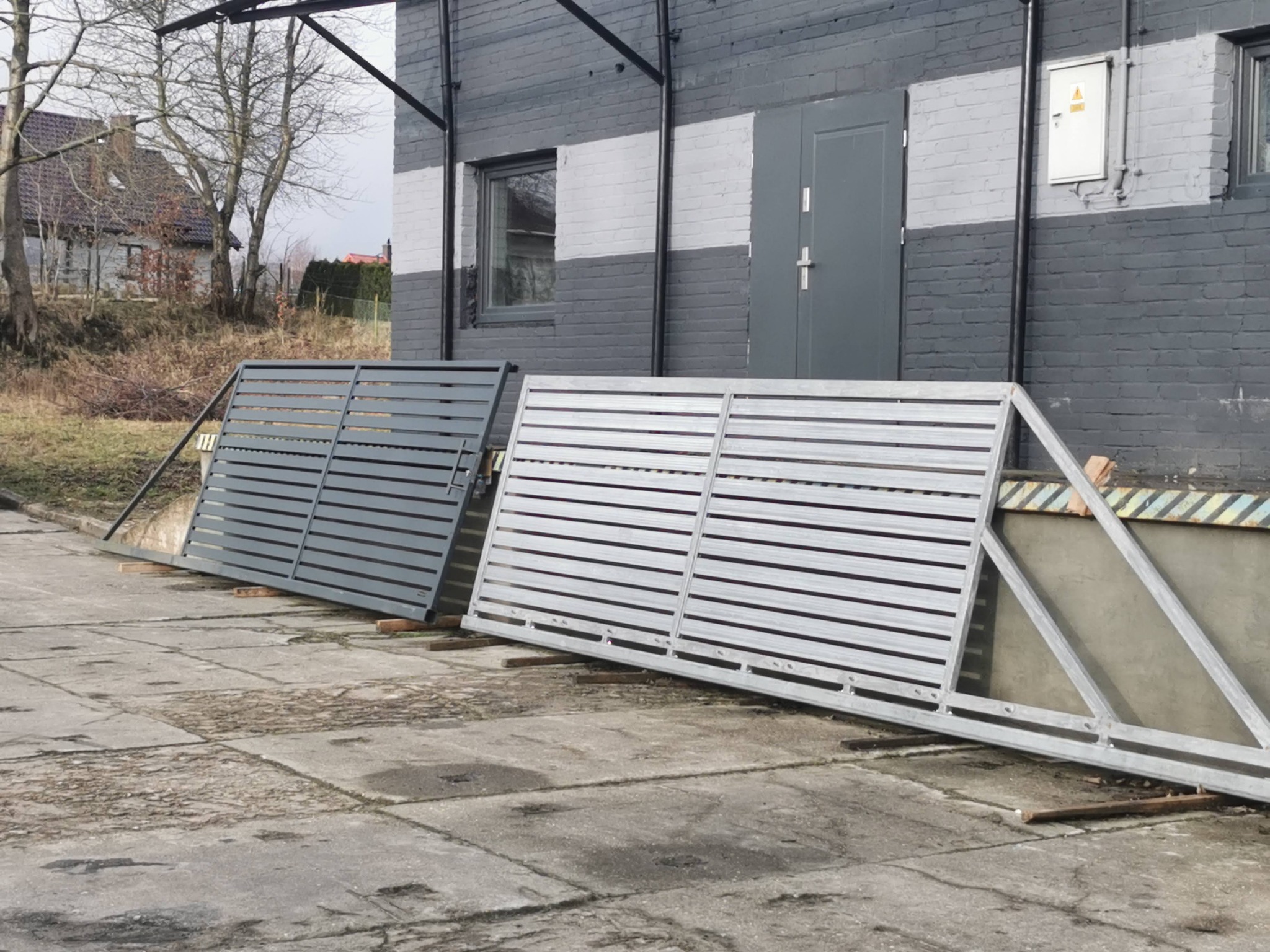 Dwie bramy przesuwne z poziomymi panelami, w odcieniach szarości, czekają na montaż w Rębiskach. Widoczne detale konstrukcyjne i surowe otoczenie.
