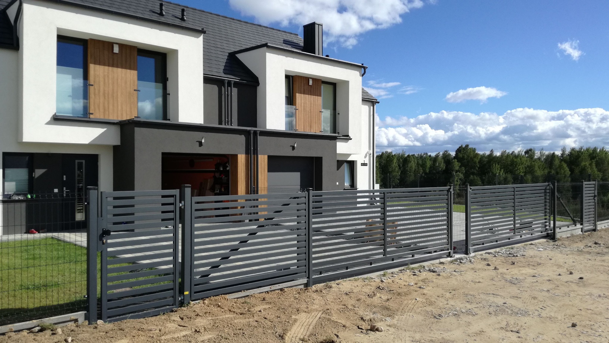 Nowoczesne ogrodzenie panelowe w kolorze grafitowym przed domem z szarą elewacją i białymi oknami. W tle błękitne niebo z chmurami i zielony las.