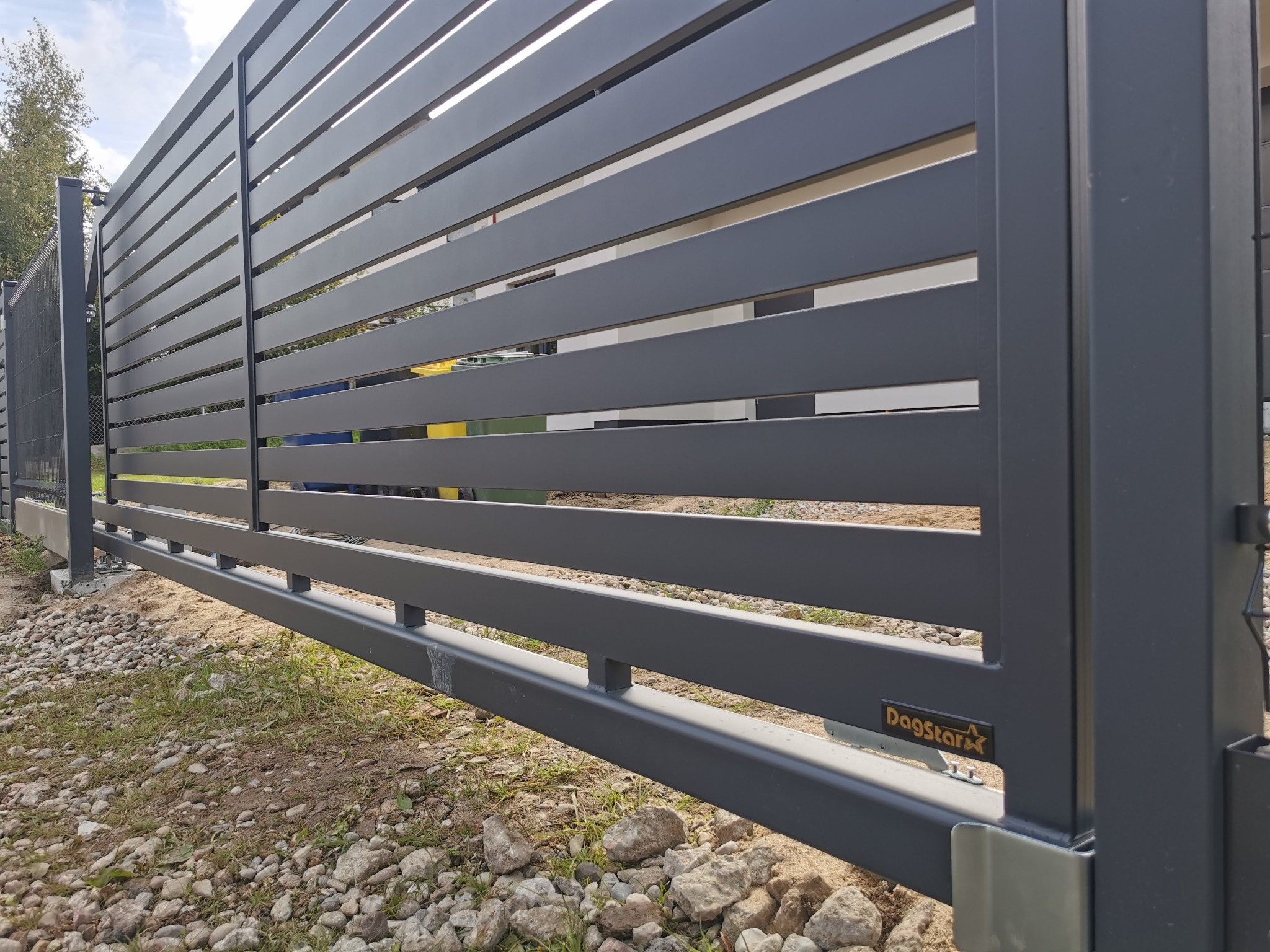Nowoczesna, przesuwna brama w kolorze grafitowym z poziomymi panelami, widoczna z bliska, z logo 'DagStar' na słupku. Tło to częściowo widoczny ogród.