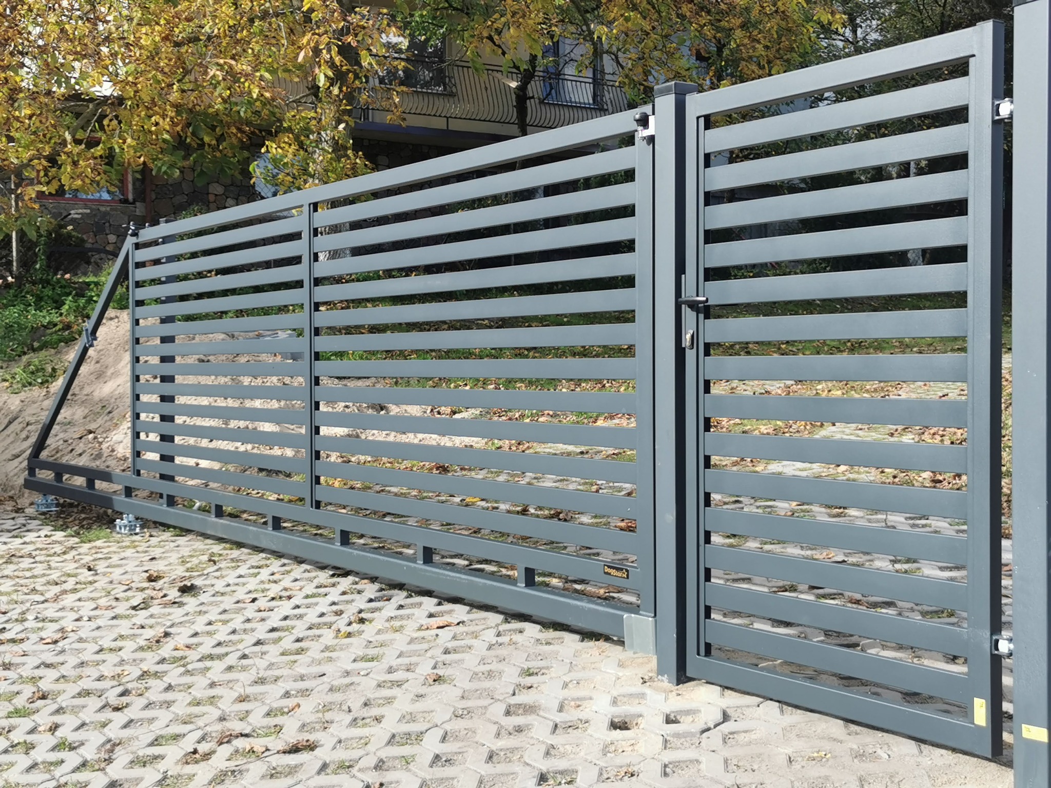 Nowoczesna, przesuwna brama metalowa w kolorze szarym z poziomymi szczeblami, zamontowana na posesji z kostką ażurową i zielenią w tle. Widoczne mocowania.