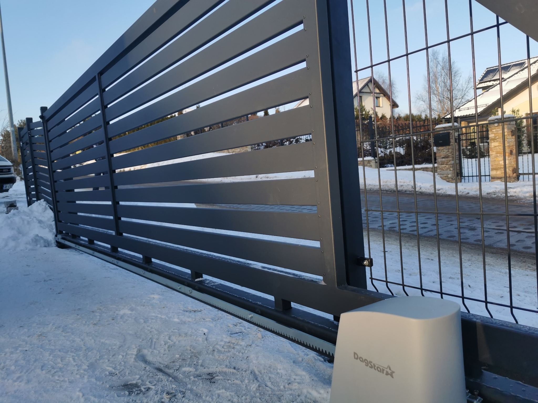 Nowoczesna, przesuwna brama wjazdowa z szarego metalu z widocznym napędem Dagstar. W tle dom jednorodzinny i siatka ogrodzeniowa. Zimowy krajobraz.