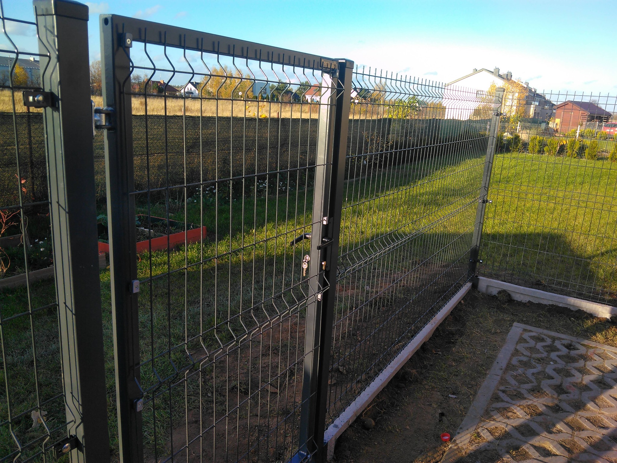 Zamknięta, metalowa furtka z panelowego ogrodzenia w kolorze grafitowym, z widoczną klamką i kluczami, na tle zielonego trawnika i zabudowań.