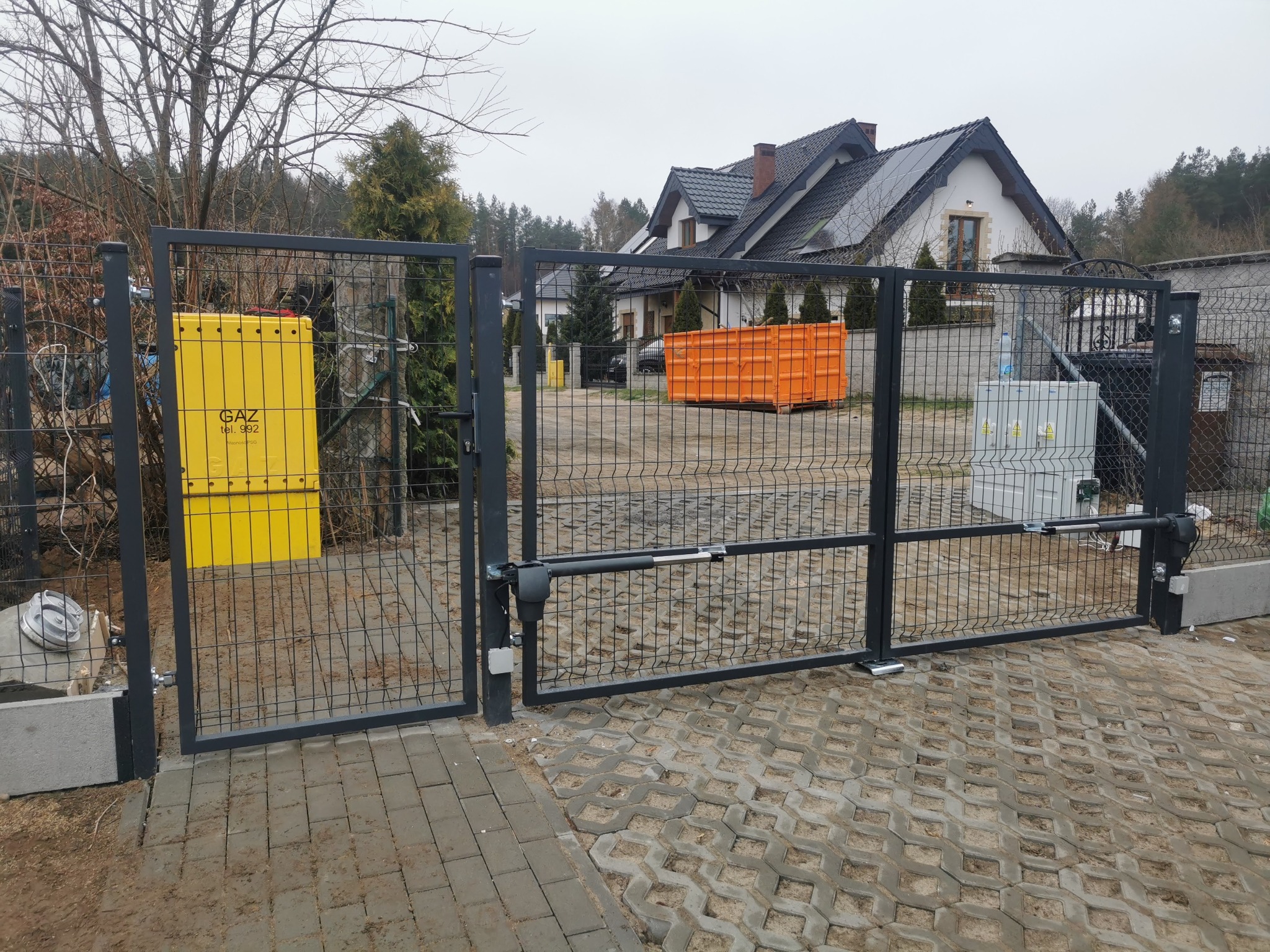 Automatyczna brama panelowa w kolorze grafitowym, zamontowana na posesji z kostką brukową. Widoczny żółty licznik gazu i dom z ciemną dachówką w tle.