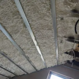 Poddasze z widoczną, jasną pianą PUR między metalowymi profilami, z fragmentami instalacji. Widoczne okno w dolnej części kadru.