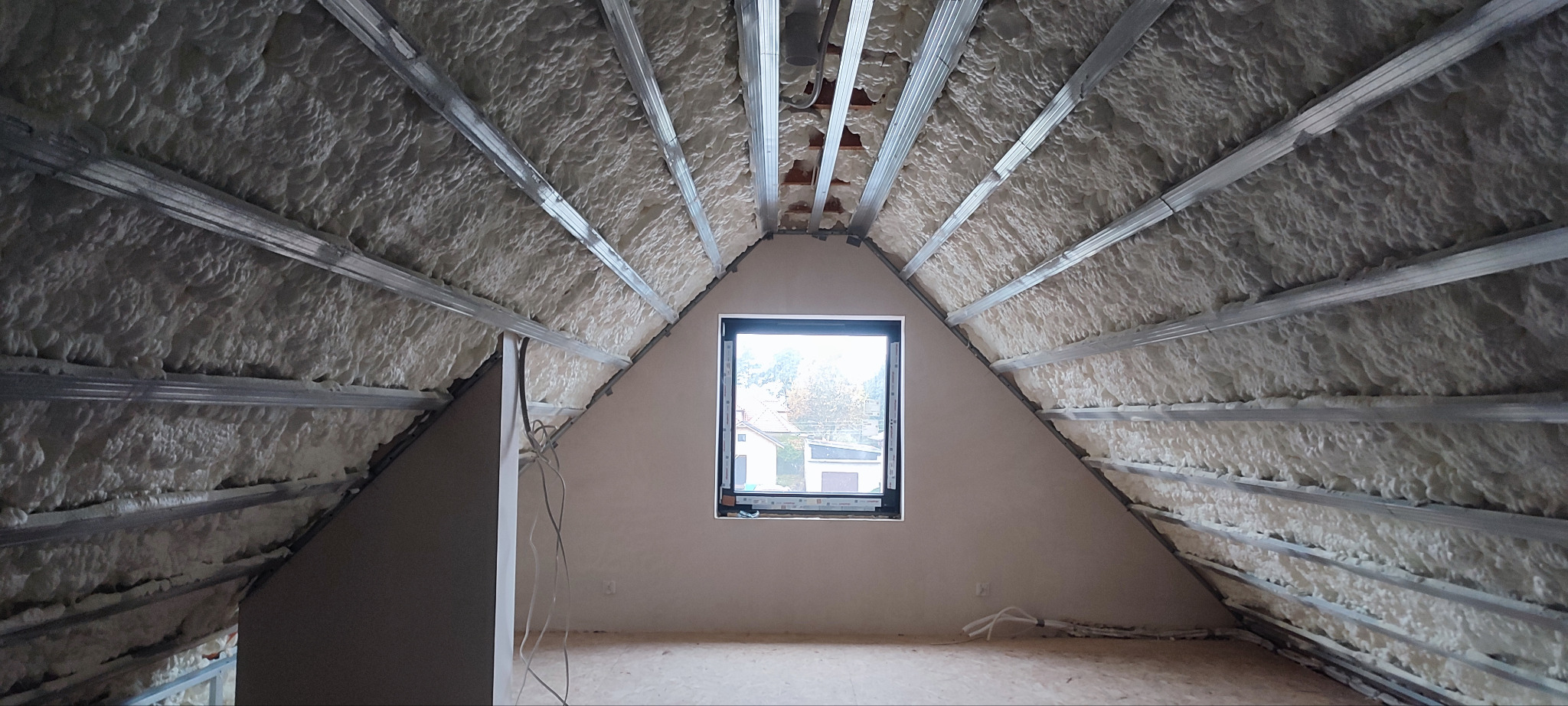 Poddasze z oknem po ociepleniu pianą PUR, widoczne metalowe profile konstrukcyjne. Piana otwartokomórkowa pokrywa skośne ściany i sufit.