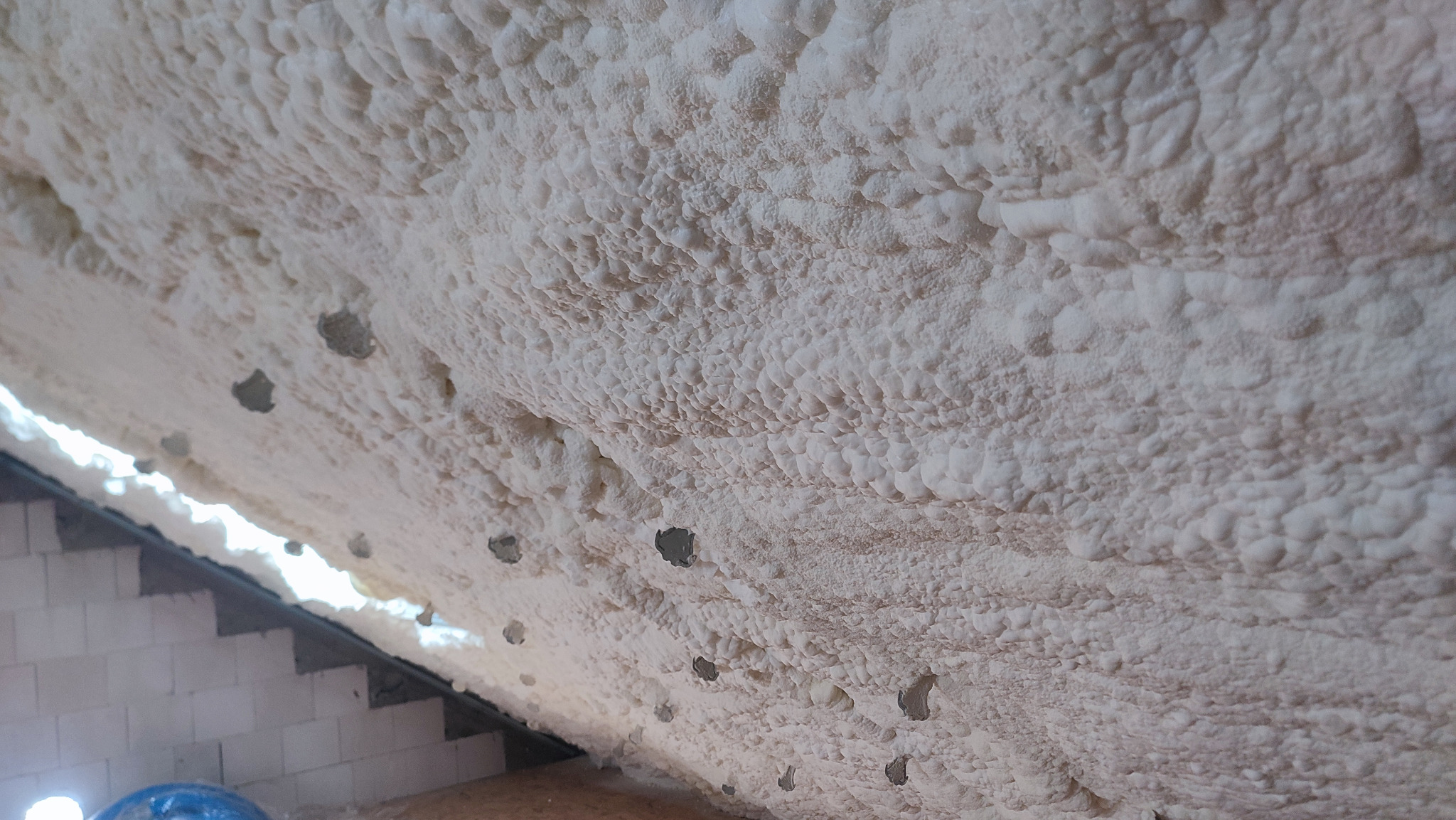 Poddasze po ociepleniu pianą PUR, widoczne liczne nierówności i charakterystyczna struktura piany. W tle ściana z pustaków i fragment schodów.