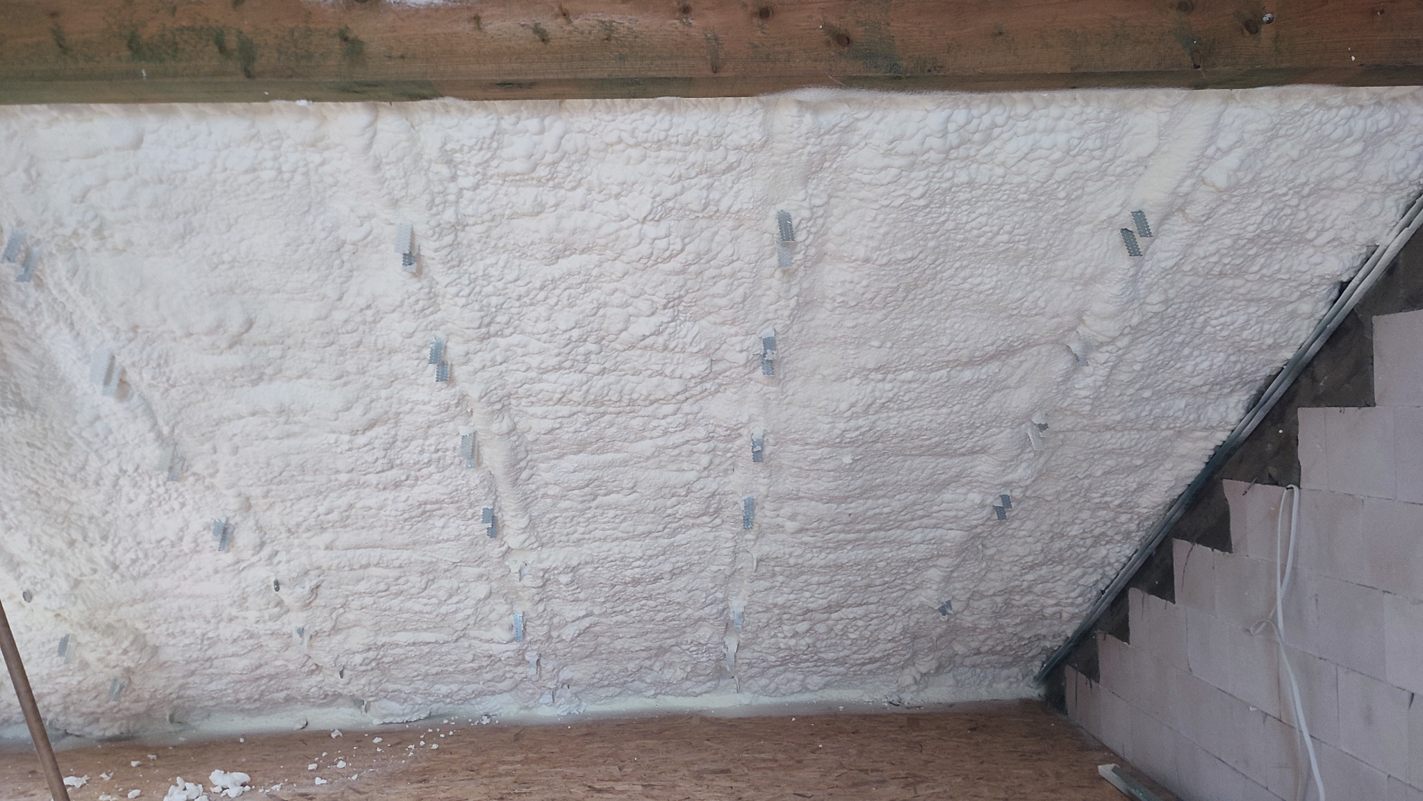 Poddasze po ociepleniu białą pianą PUR, widoczne metalowe elementy mocujące. Fragment ściany z bloczków po prawej stronie. Oświetlenie naturalne.