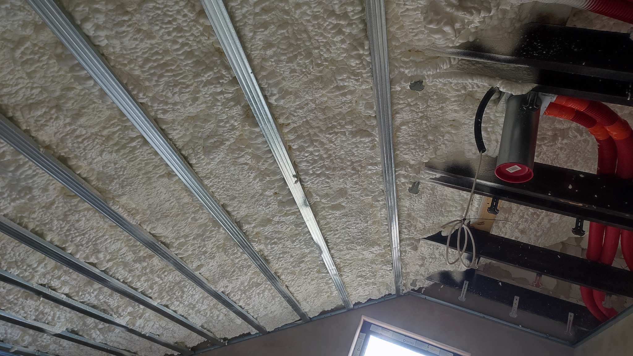 Poddasze z widoczną, jasną pianą PUR między metalowymi profilami, z fragmentami instalacji. Widoczne okno w dolnej części kadru.