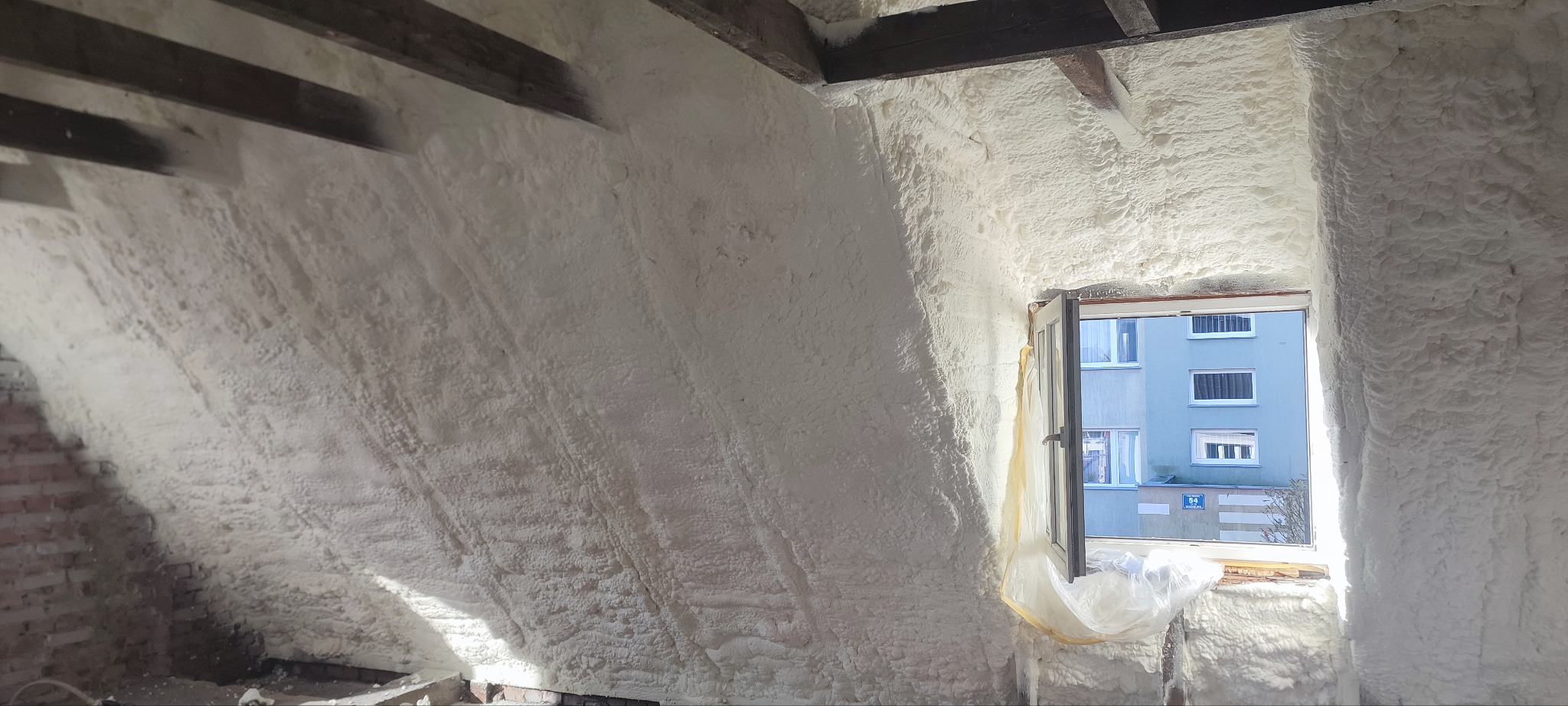 Poddasze po ociepleniu pianą PUR otwartokomórkową, widoczne krokwie i okno z widokiem na budynek. Izolacja SmartPur na skośnych ścianach i suficie.