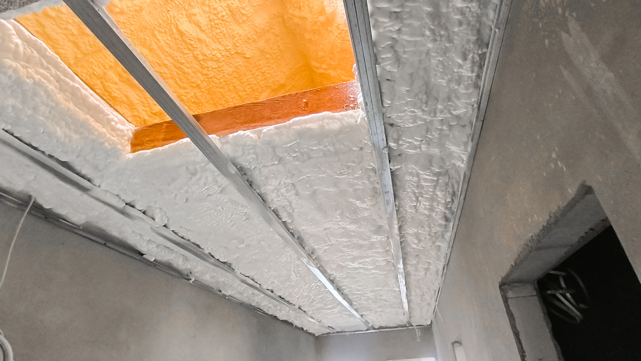 Poddasze z widoczną strukturą piany PUR po natrysku, z metalowymi profilami. Fragment pomarańczowej membrany widoczny w przestrzeni między krokwiami.