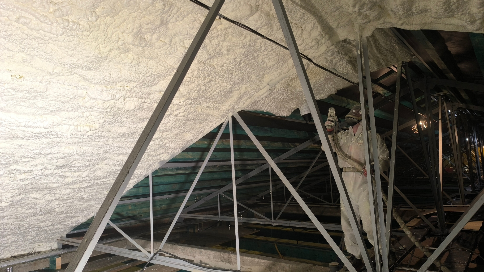 Aplikacja pianki PUR na poddaszu z widoczną konstrukcją dachu i pracownikiem w kombinezonie ochronnym, wykonującym natrysk izolacji. Oświetlenie robocze.