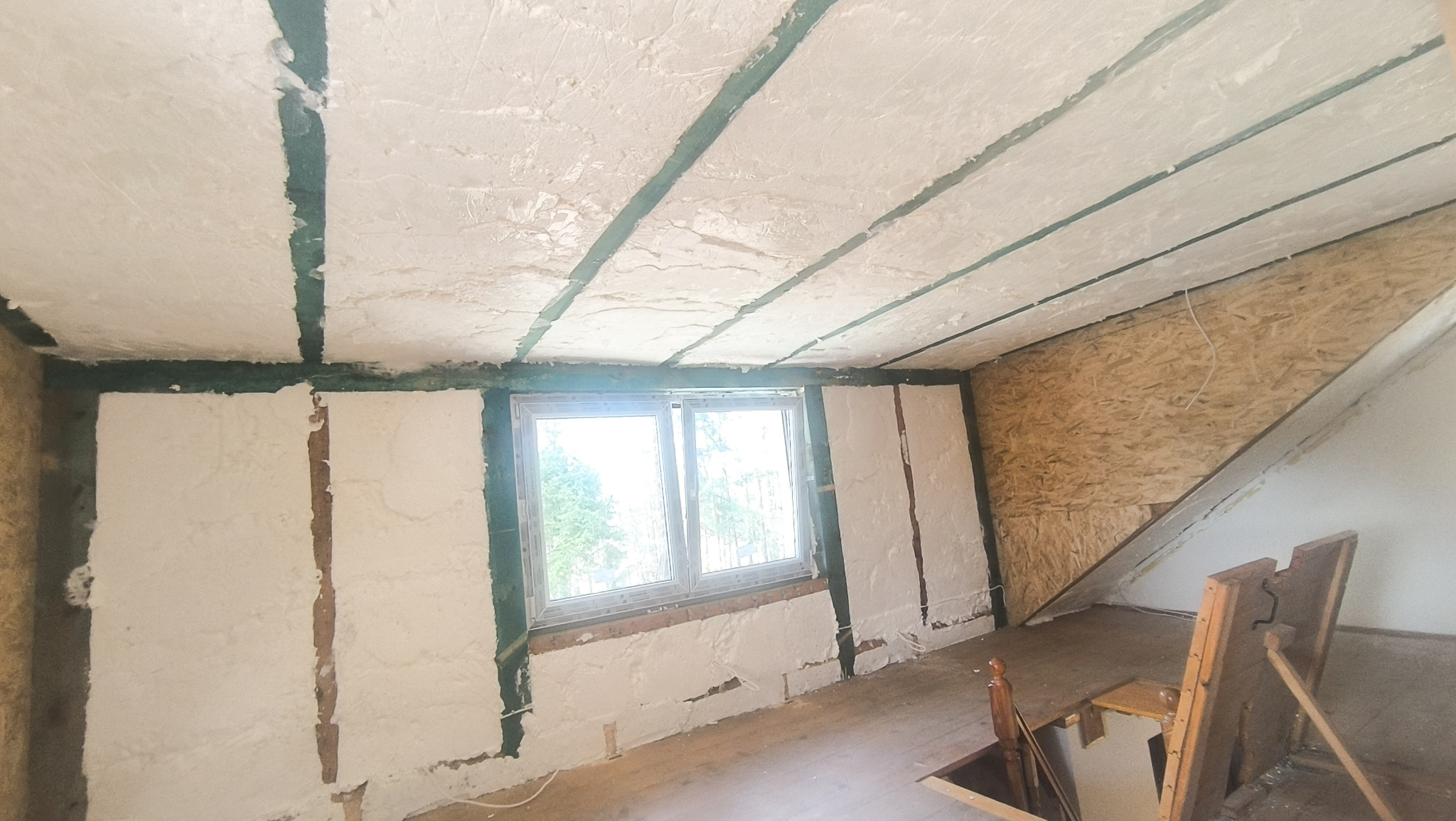 Poddasze po ociepleniu pianą PUR otwartokomórkową, widoczne zielone elementy konstrukcyjne, okno i fragment schodów. Ocieplenie wykonane metodą natryskową.