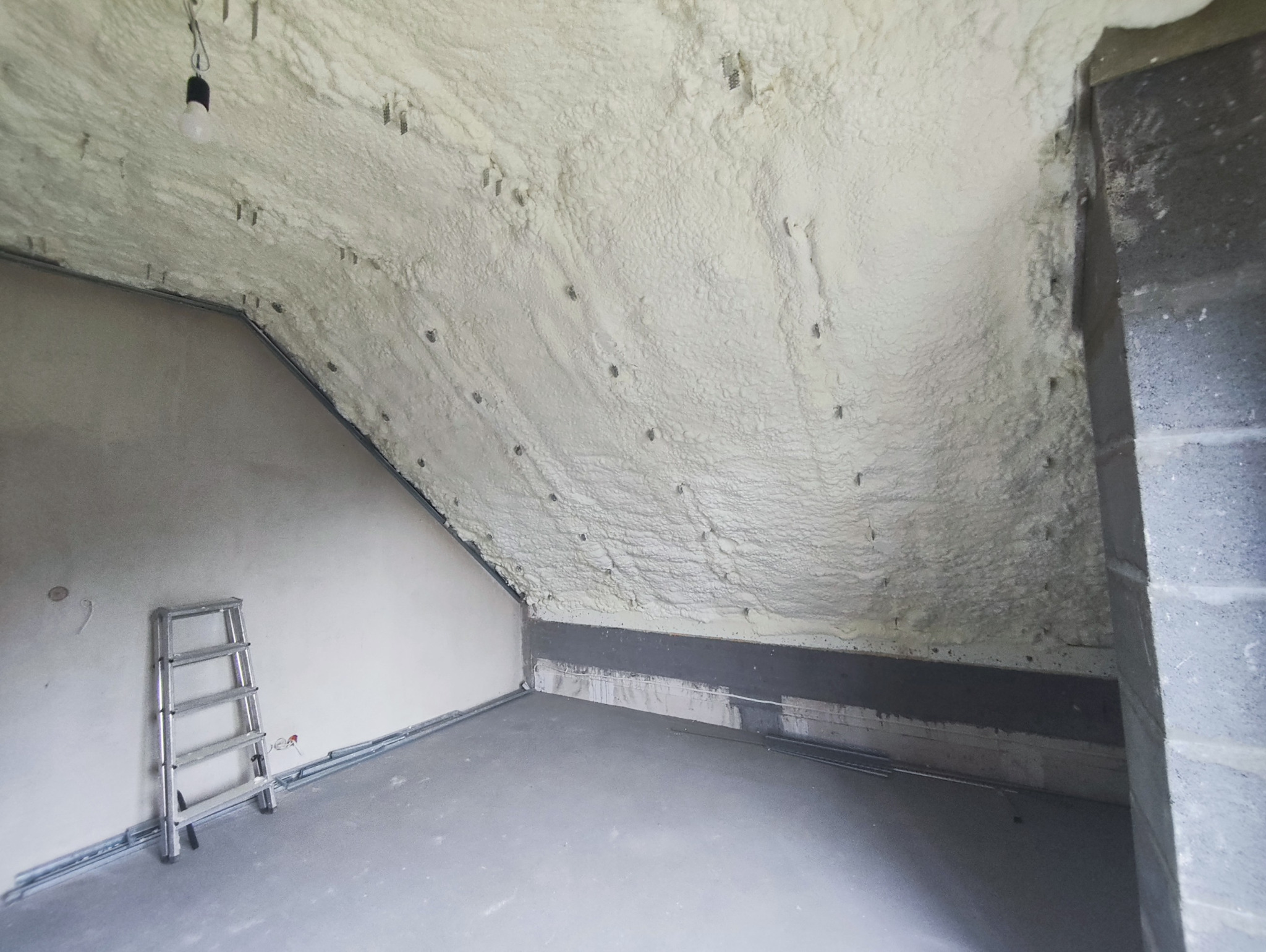 Poddasze po ociepleniu pianką PUR otwartokomórkową. Widoczna drabina oparta o ścianę, sufit pokryty pianą, wystające elementy montażowe. Oświetlenie żarówką.