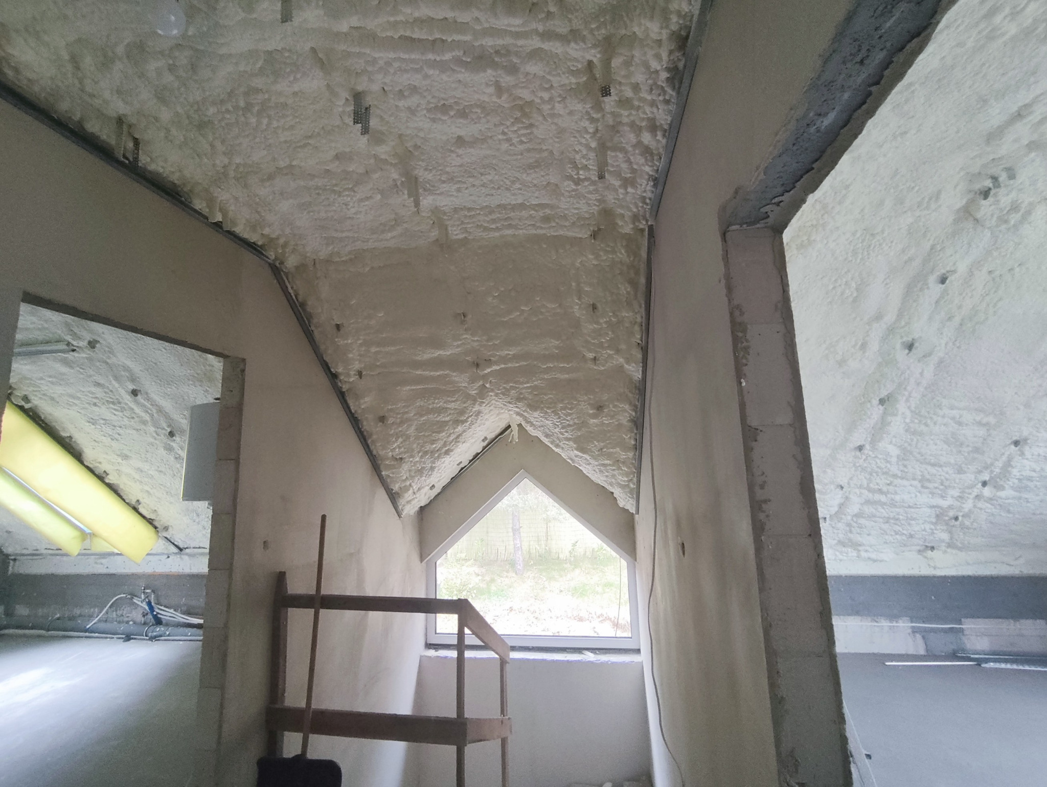 Poddasze po ociepleniu pianą PUR otwartokomórkową, widoczne krokwie i okno dachowe. Biała, porowata struktura piany pokrywa skośne powierzchnie.