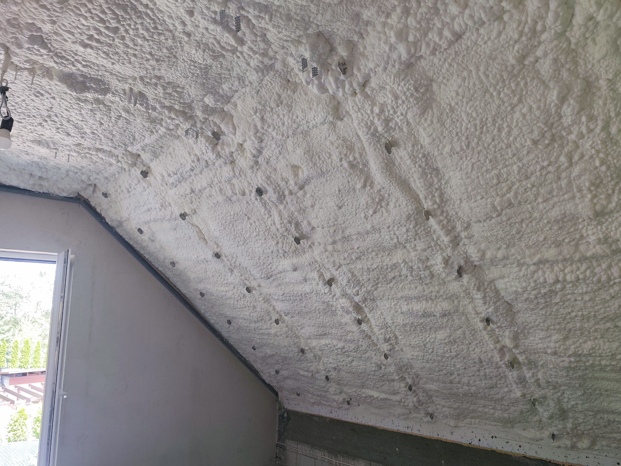 Poddasze po ociepleniu pianą PUR otwartokomórkową, widoczne metalowe mocowania, fragment okna i ściany. Tekstura piany przypomina gęstą, białą watę.