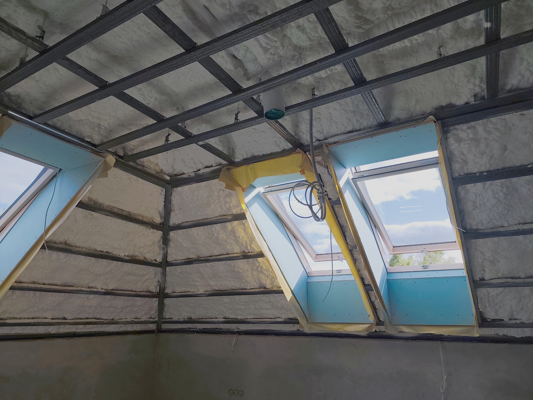 Poddasze z oknami dachowymi w trakcie ocieplania pianą PUR, widoczna metalowa konstrukcja i zabezpieczone okna folią. Etap prac przed wykończeniem.