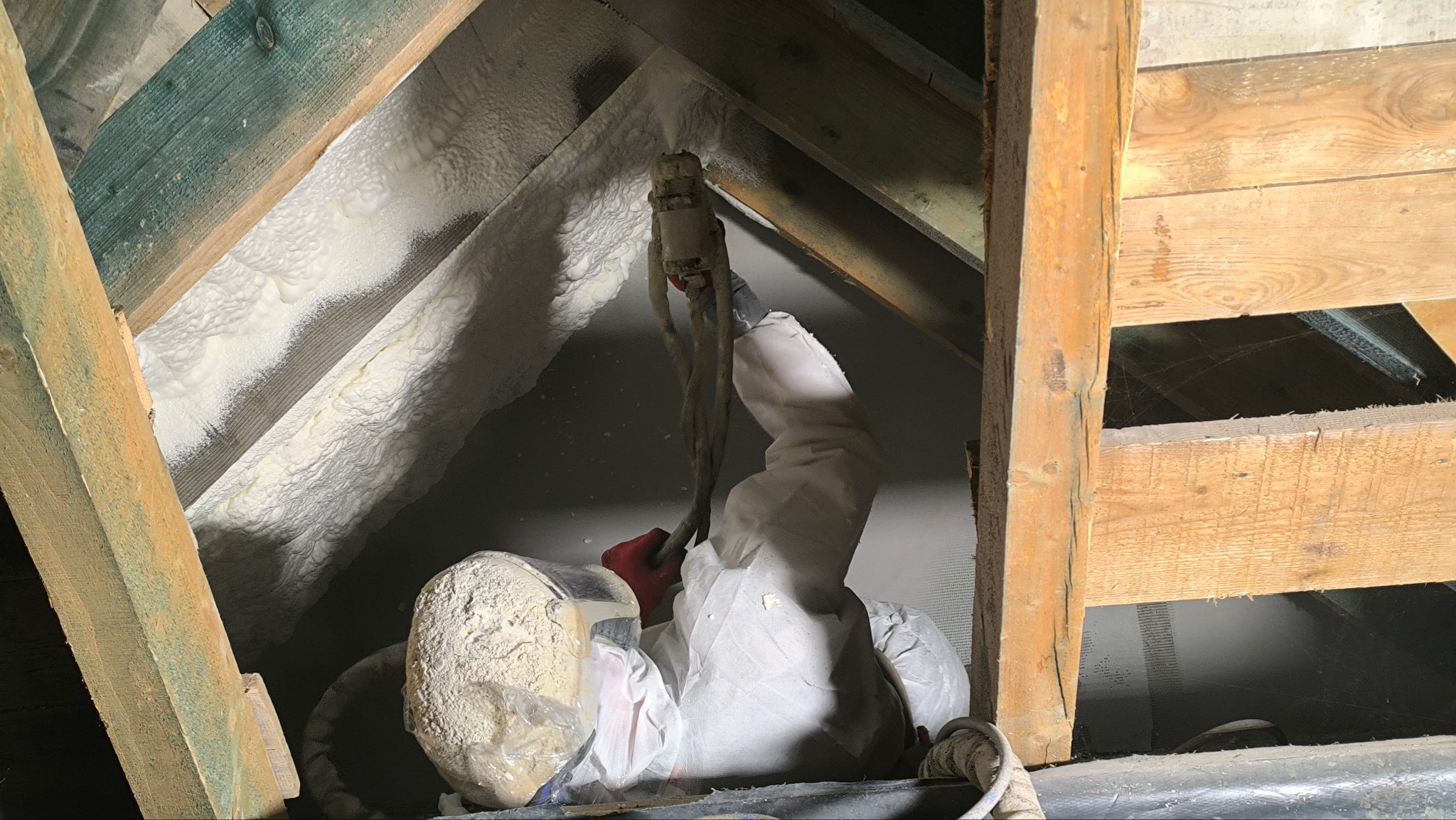 Aplikacja pianki PUR w przestrzeni poddasza przez pracownika w kombinezonie ochronnym. Widoczne drewniane elementy konstrukcyjne i świeżo nałożona, jasna pianka.
