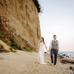 Robert Malkusz - Para młoda, boso, spaceruje po piaszczystej plaży trzymając się za ręce. W tle klif i morze. Ujęcie z sesji ślubnej, naturalne światło.