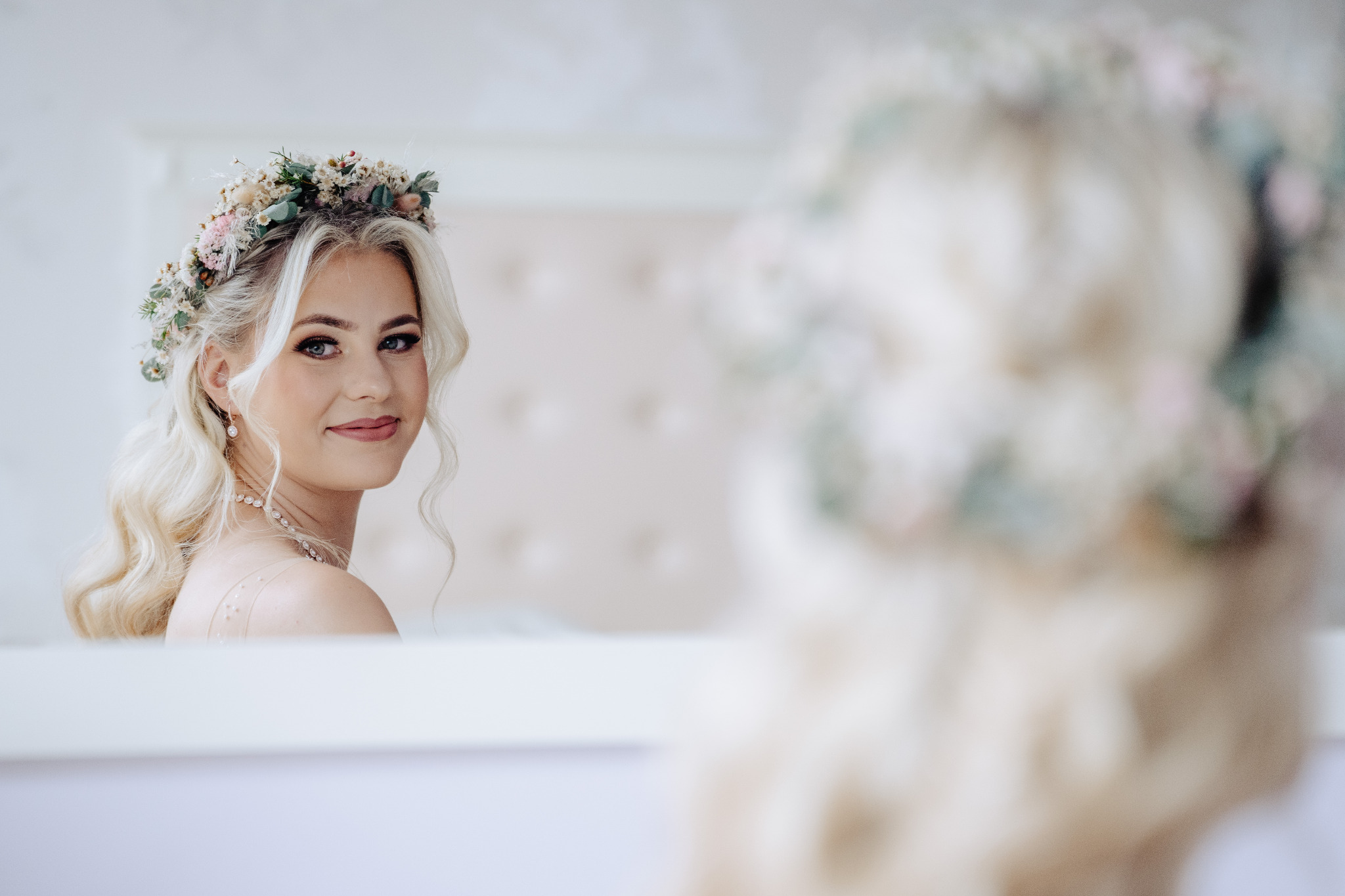 Portret panny młodej z wiankiem, delikatny makijaż, blond włosy w stylu retro, uśmiech, w tle rozmyty bukiet ślubny. Subtelne oświetlenie, jasna kolorystyka.