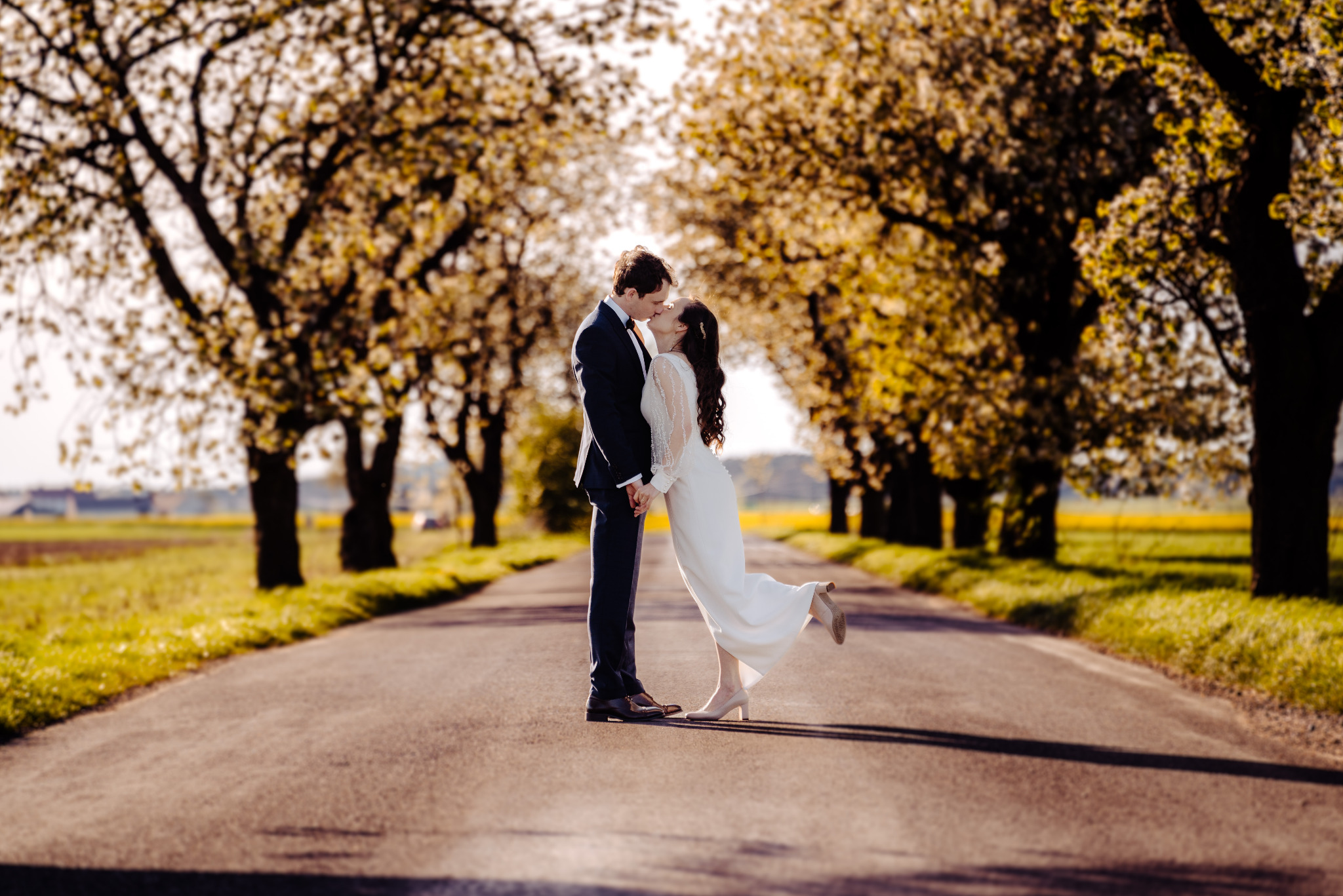 Para młoda w objęciach na drodze otoczonej kwitnącymi drzewami. Panna młoda w białej sukni unosi nogę, tworząc dynamiczną pozę. Romantyczna sceneria, ciepłe światło.