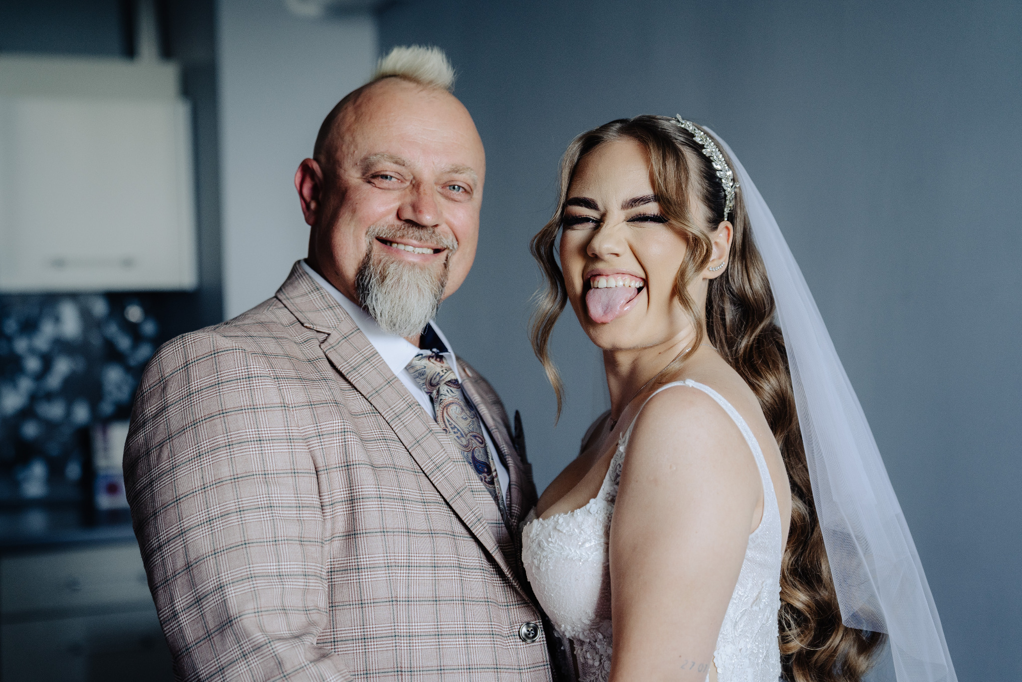 Uśmiechnięty pan młody w kraciastej marynarce i panna młoda z wyciągniętym językiem, w tiarze i welonie, pozują do zdjęcia w pomieszczeniu.