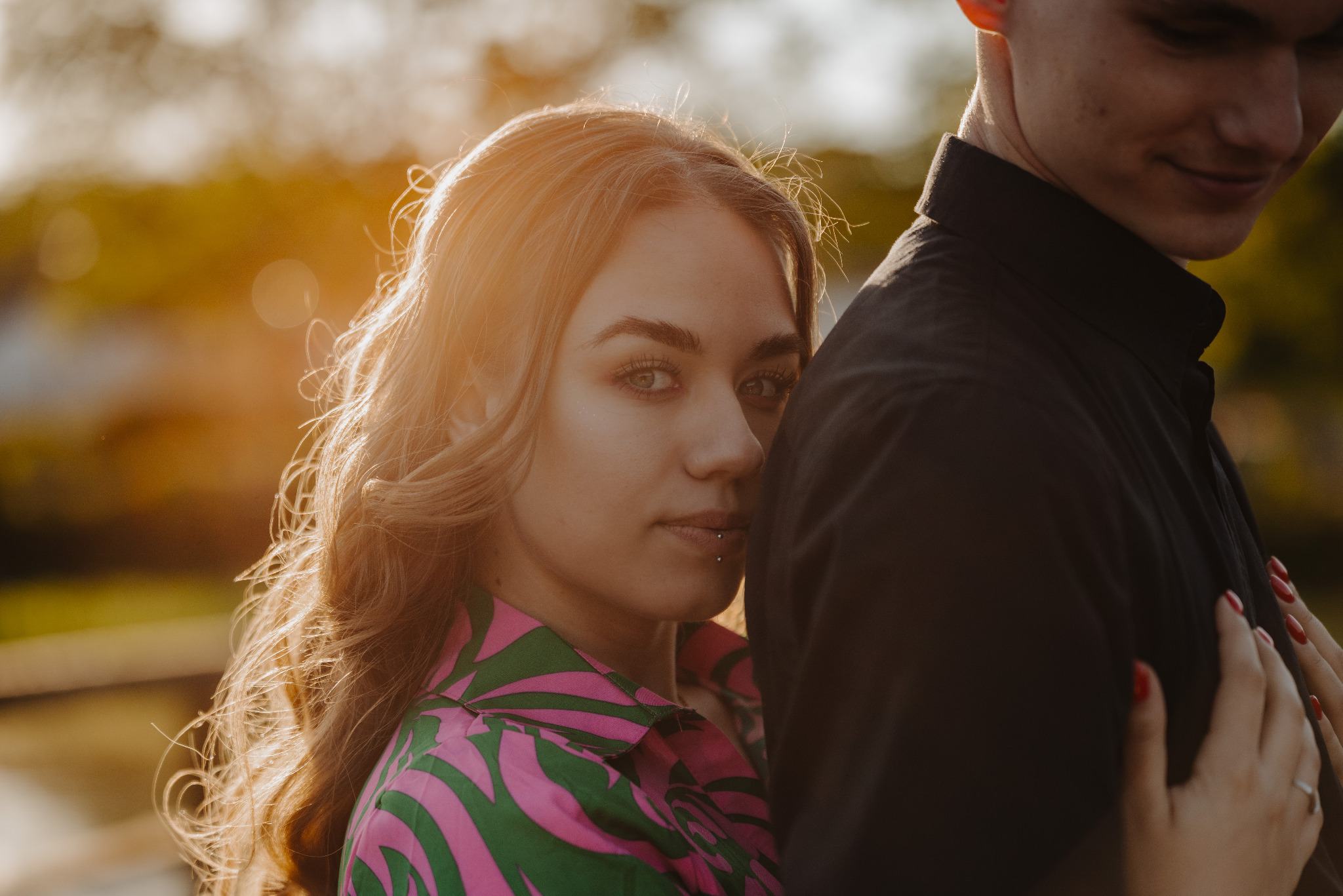 Portret kobiety z piercingiem w wardze, opierającej się o mężczyznę w czarnej koszuli, w ciepłym, naturalnym świetle. Romantyczna atmosfera.