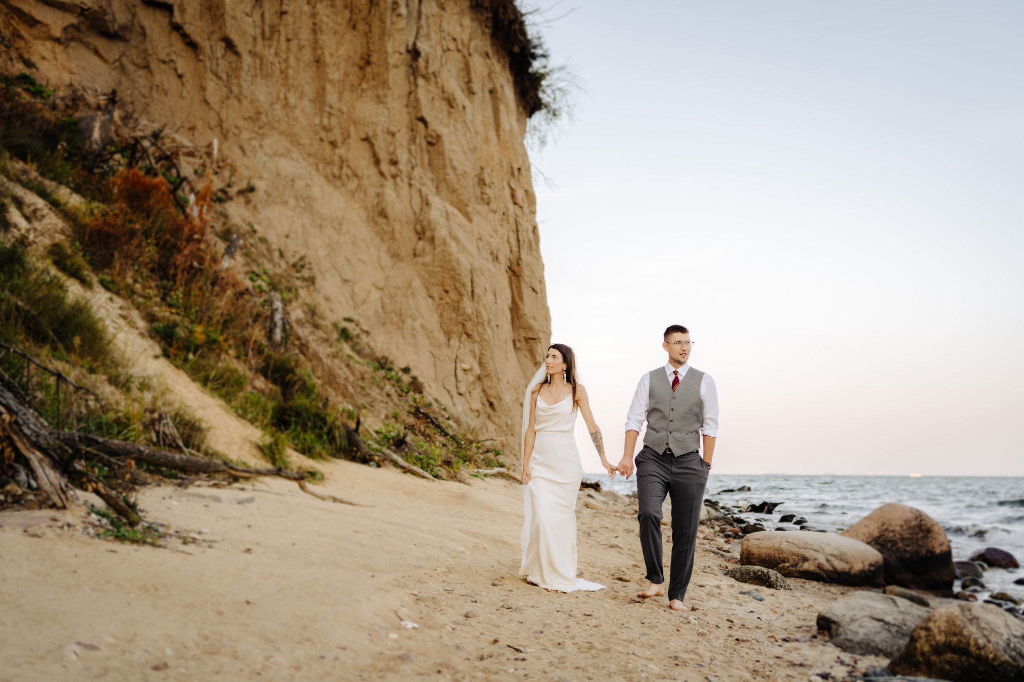 Para młoda, boso, spaceruje po piaszczystej plaży trzymając się za ręce. W tle klif i morze. Ujęcie z sesji ślubnej, naturalne światło.
