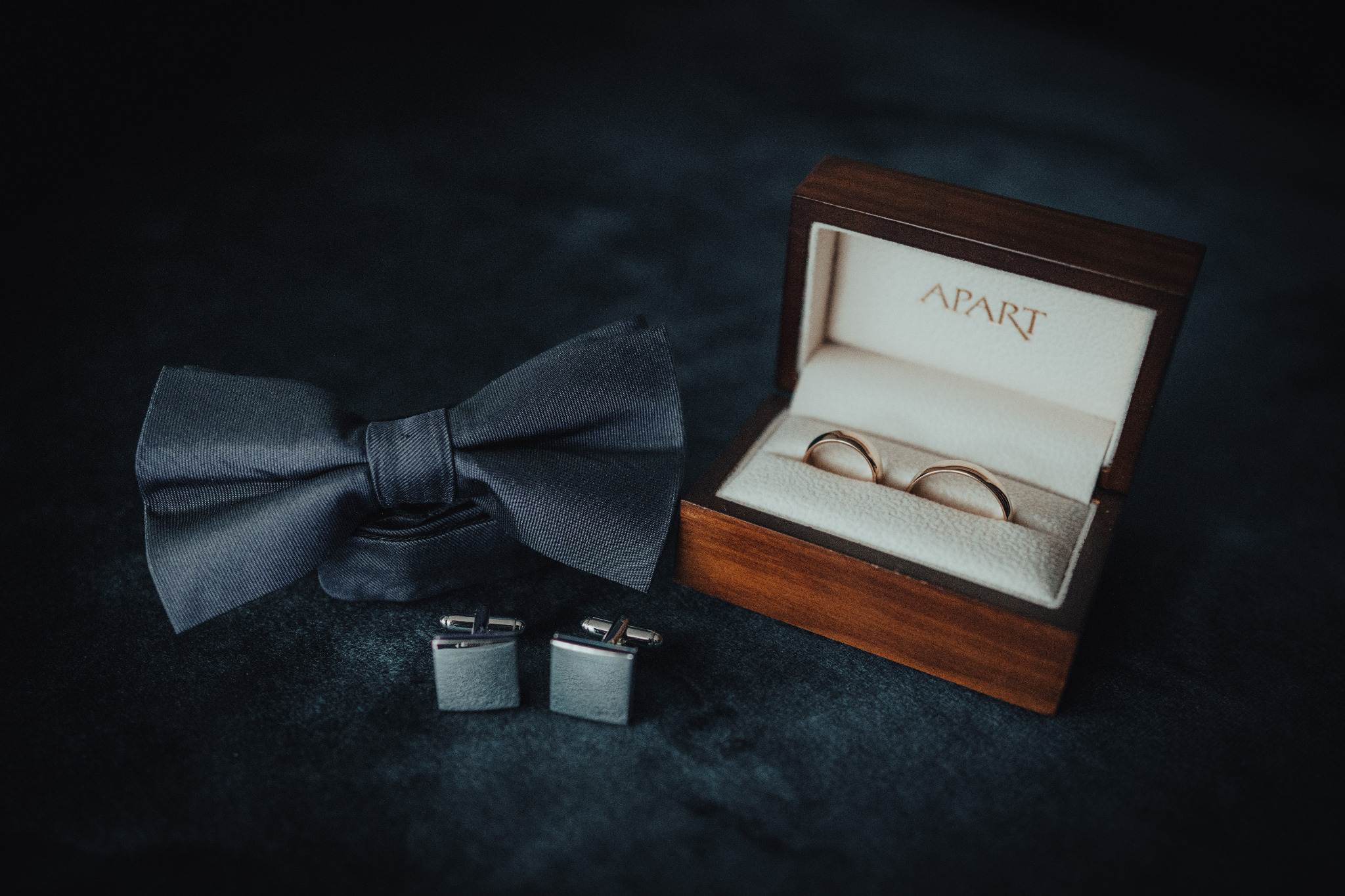Elegancki zestaw ślubny: granatowa mucha, srebrne spinki do mankietów i obrączki w drewnianym pudełku marki Apart na ciemnym tle. Subtelne światło podkreśla detale.