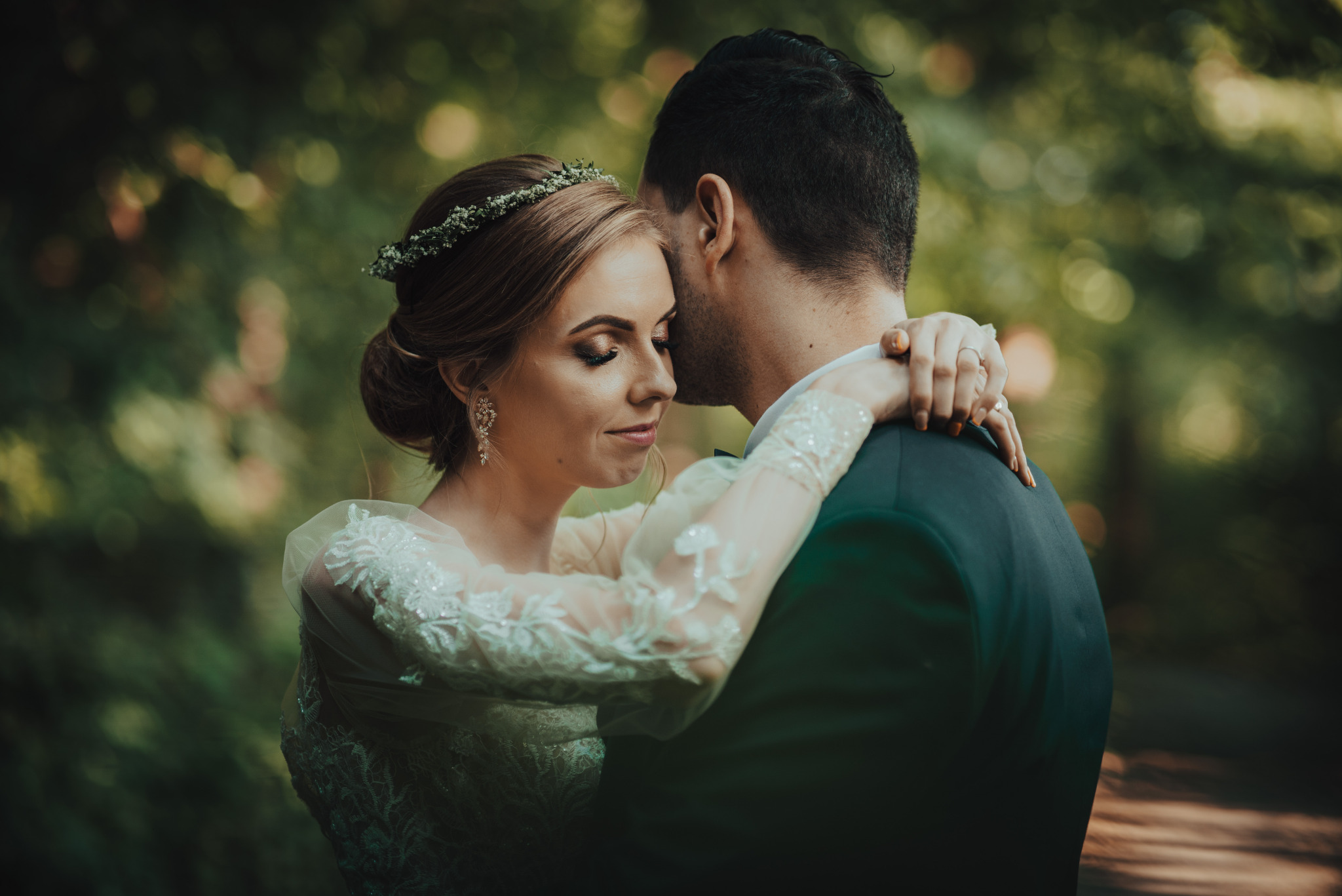 Uśmiechnięta panna młoda w koronie z kwiatów, obejmowana przez pana młodego w ciemnym garniturze na tle rozmytej zieleni.