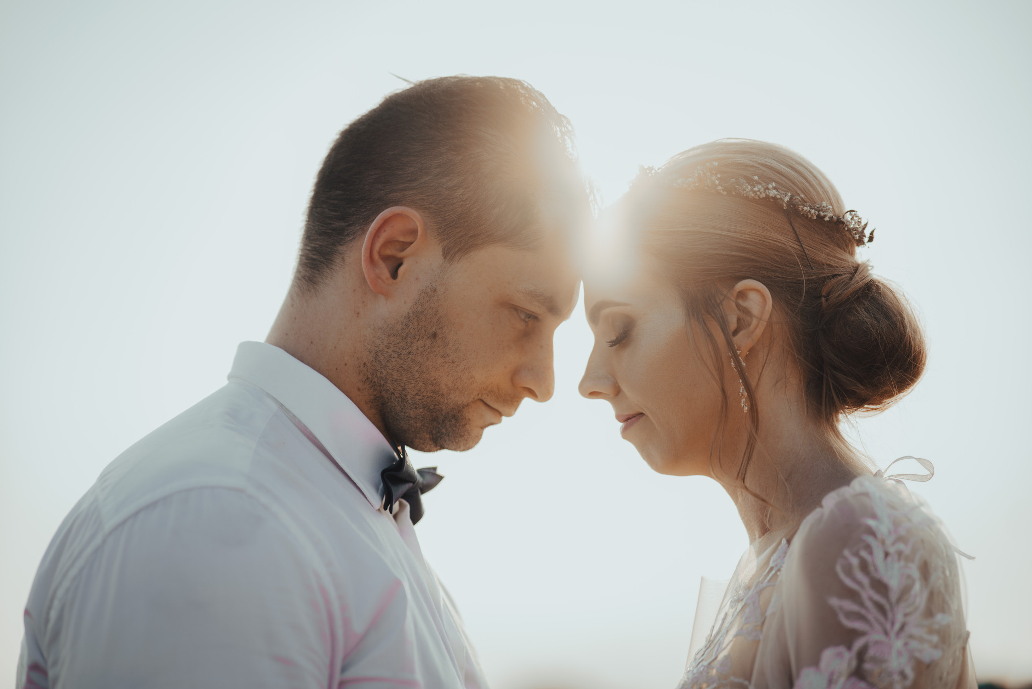 Delikatne zbliżenie profilu pary młodej, czoła dotykają się w czułym geście. Ona w sukni z koronką i wiankiem, on w białej koszuli z muchą. Ciepłe, rozproszone światło.