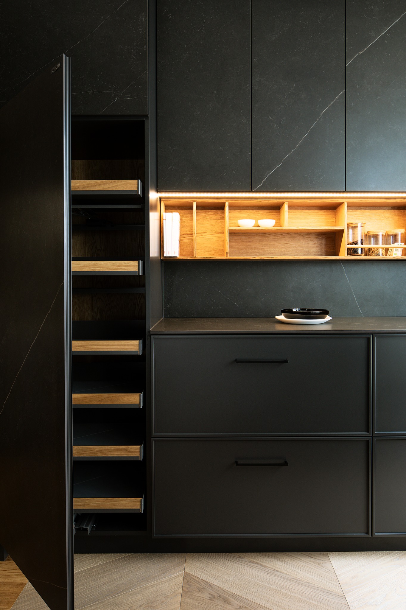 Nowoczesna, czarna zabudowa kuchenna z drewnianymi akcentami i subtelnym oświetleniem LED. Minimalistyczny design z blatem imitującym kamień i otwartymi półkami.