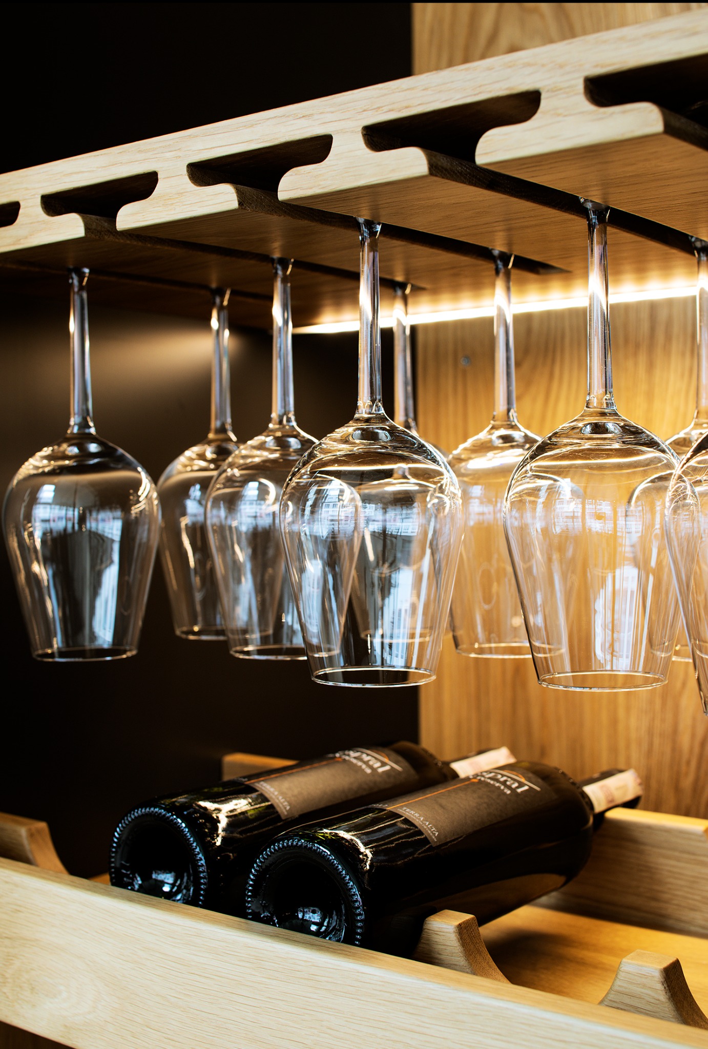Drewniany regał na wino z podwieszonymi kieliszkami, dwie butelki wina na dolnej półce, ciepłe oświetlenie, nowoczesny design.