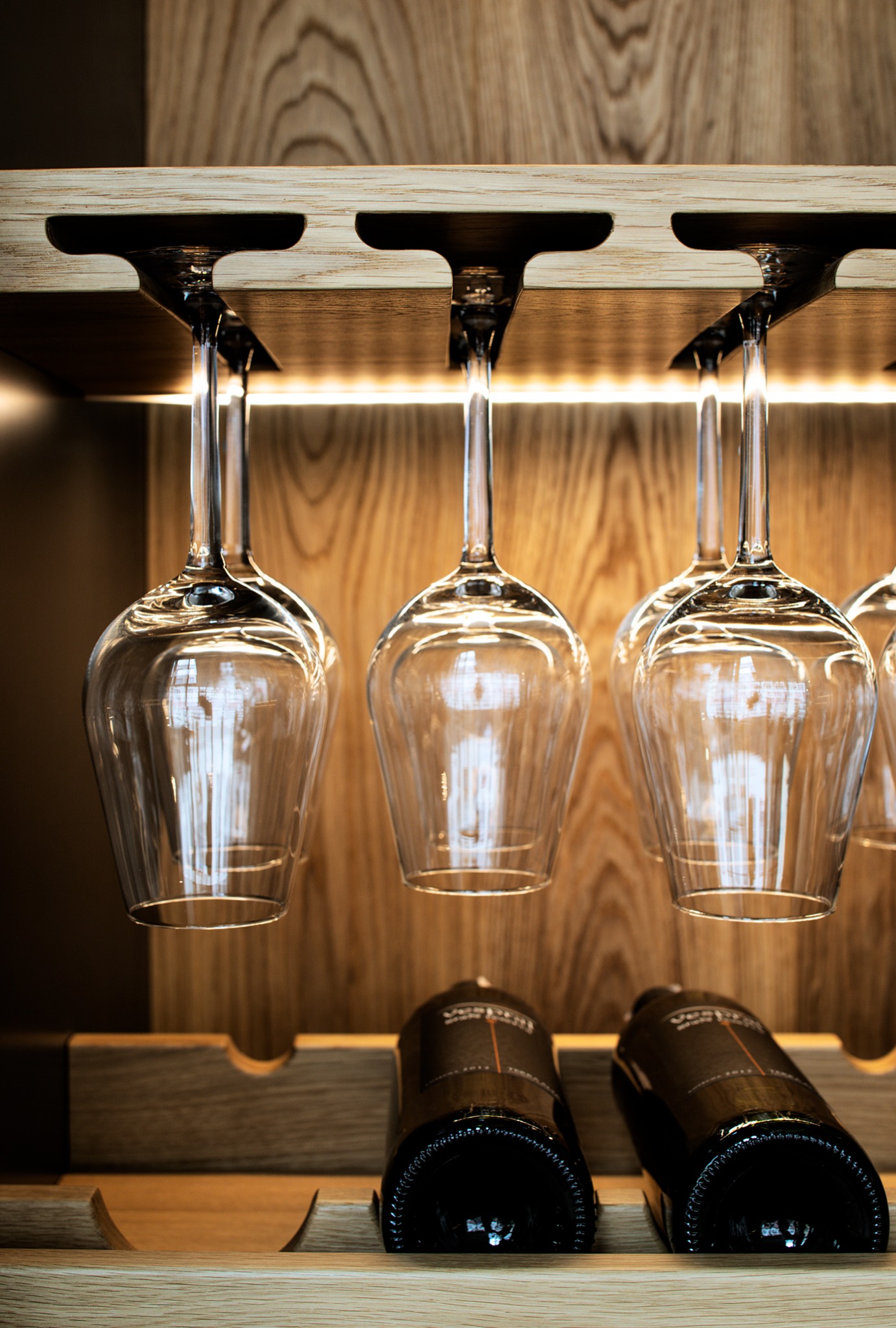 Drewniany barek z podświetleniem LED, kieliszki do wina wiszące do góry nogami, dwie butelki wina leżące na półce. Nowoczesny design.