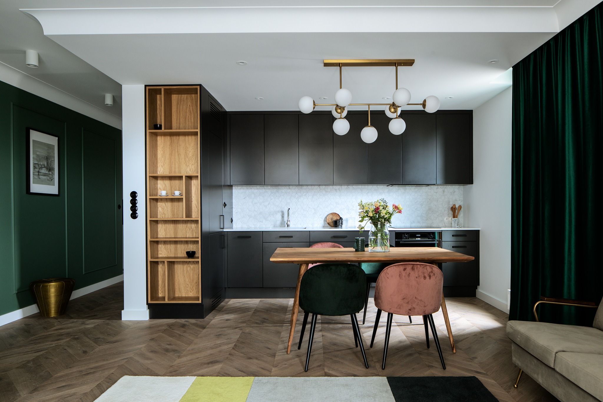 Nowoczesna kuchnia z jadalnią: ciemne szafki, drewniany stół z welurowymi krzesłami w odcieniach zieleni i różu, parkiet w jodełkę, designerska lampa i zielone zasłony.