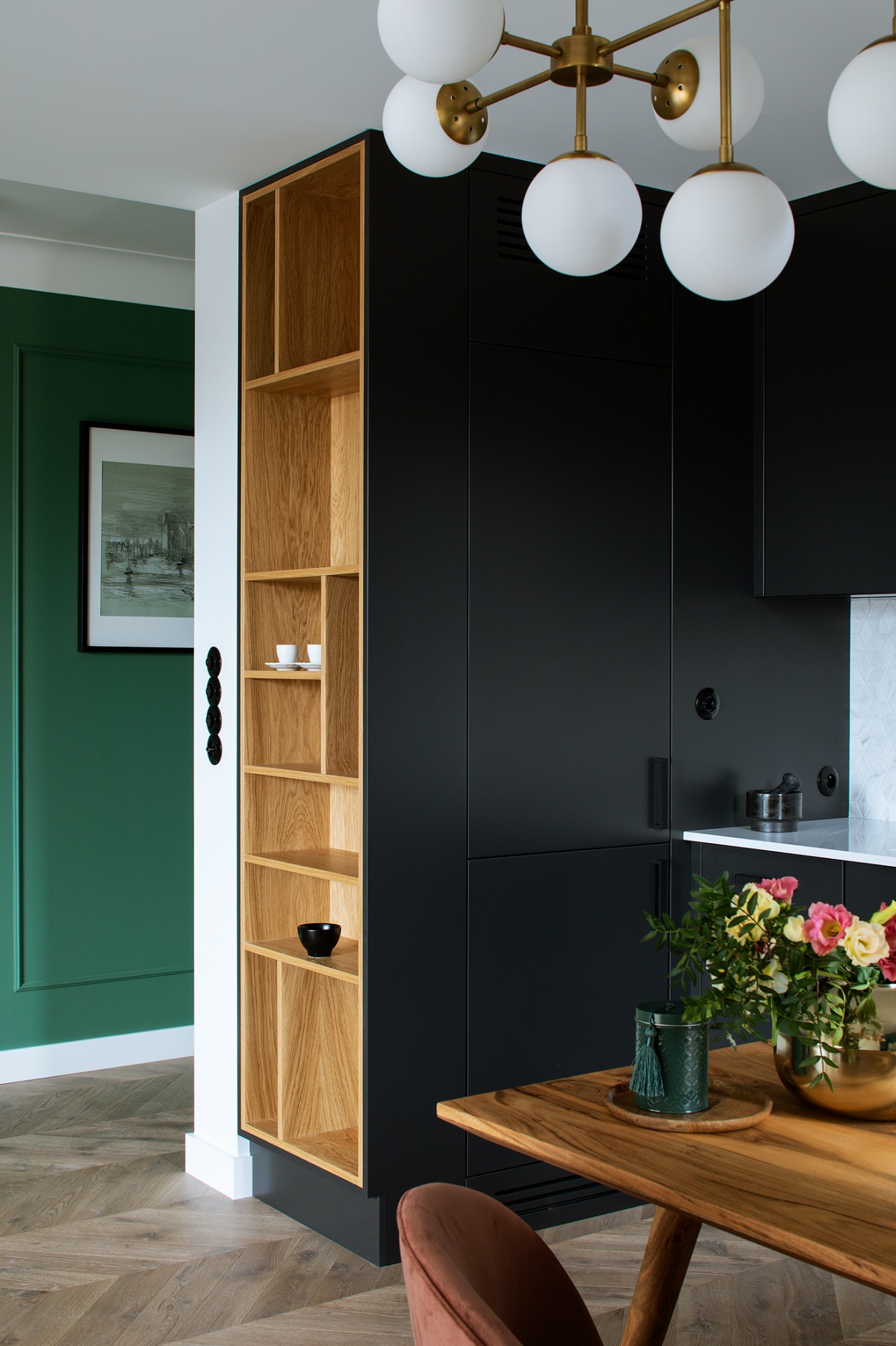 Nowoczesna kuchnia z czarnymi szafkami, drewnianym regałem i blatem. Wazon ze świeżymi kwiatami na stole, parkiet w jodełkę i designerska lampa sufitowa.