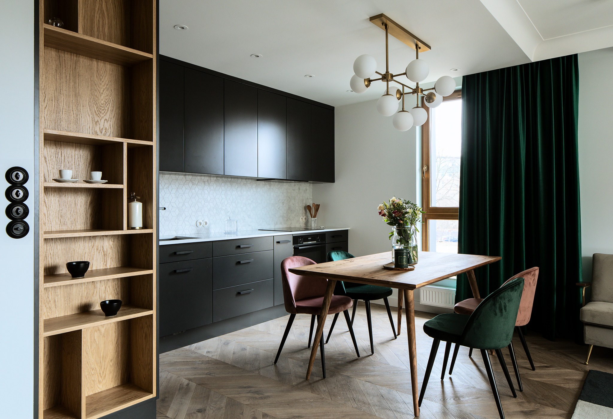 Stylowa jadalnia z drewnianym stołem i krzesłami w odcieniach różu i zieleni. W tle ciemna zabudowa kuchenna, parkiet w jodełkę i zielone zasłony.