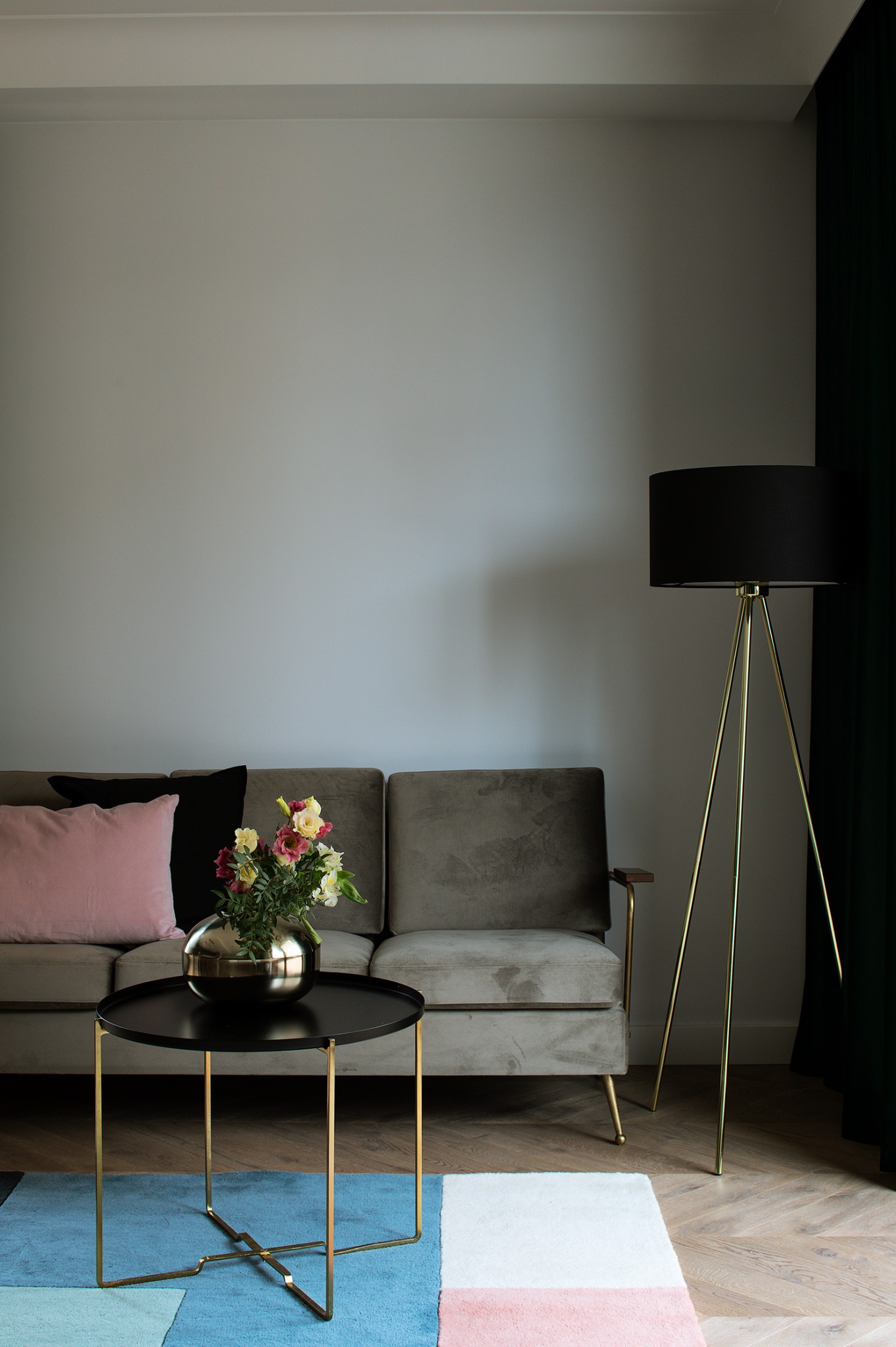 Stylowy salon z szarą sofą, kwiatami w złotej kuli na czarnym stoliku i lampą na złotym statywie. Kolorowy dywan i ciemnozielone zasłony dopełniają aranżację.